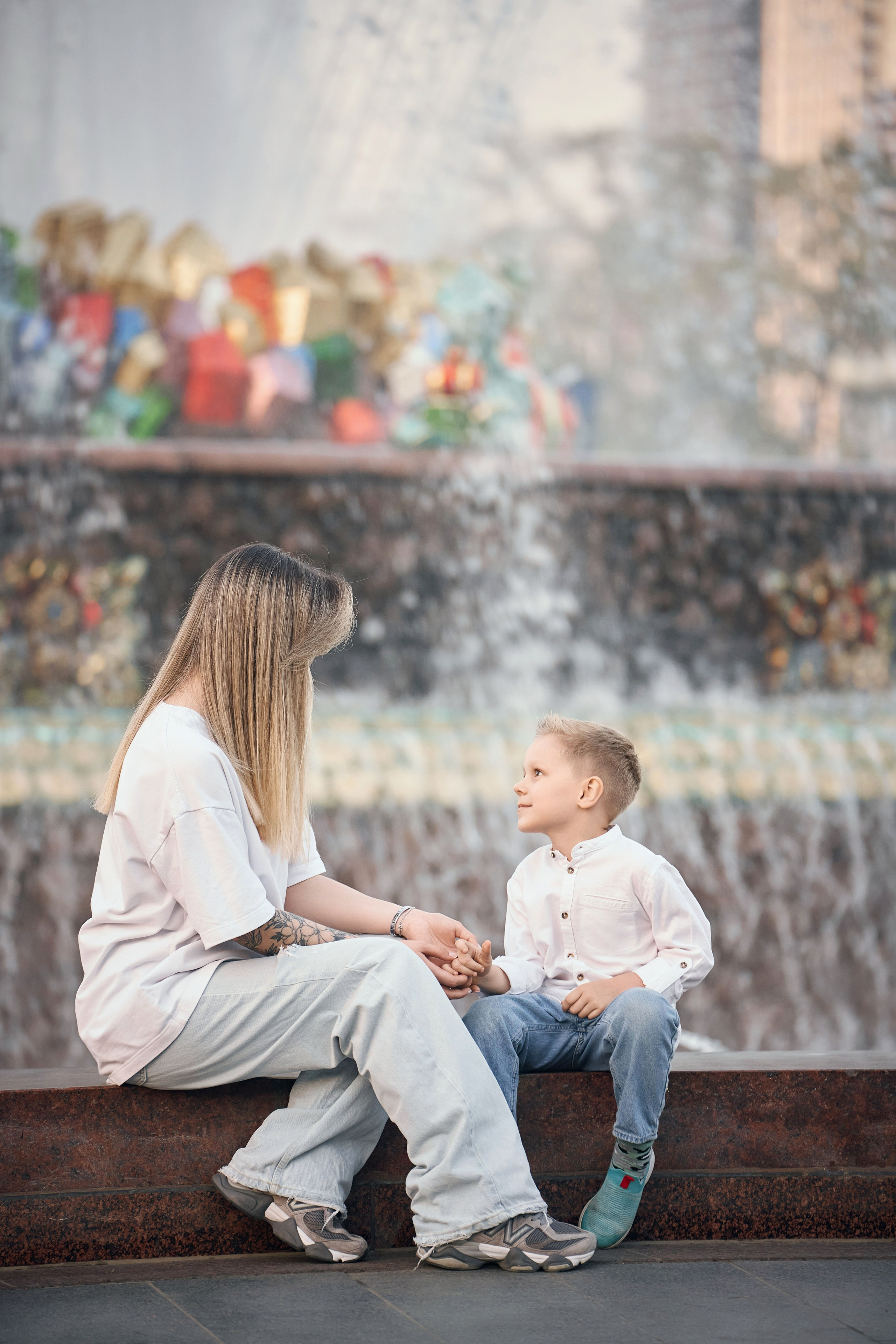 кадр с семейной фотосессии у фонтана каменный цветок, мама и сын держаться за руки, мама одета в белую рубашку и голубые джинсы, сын одет в белую рубашку и синие джинсы, фотосесссия в голубой час после заката