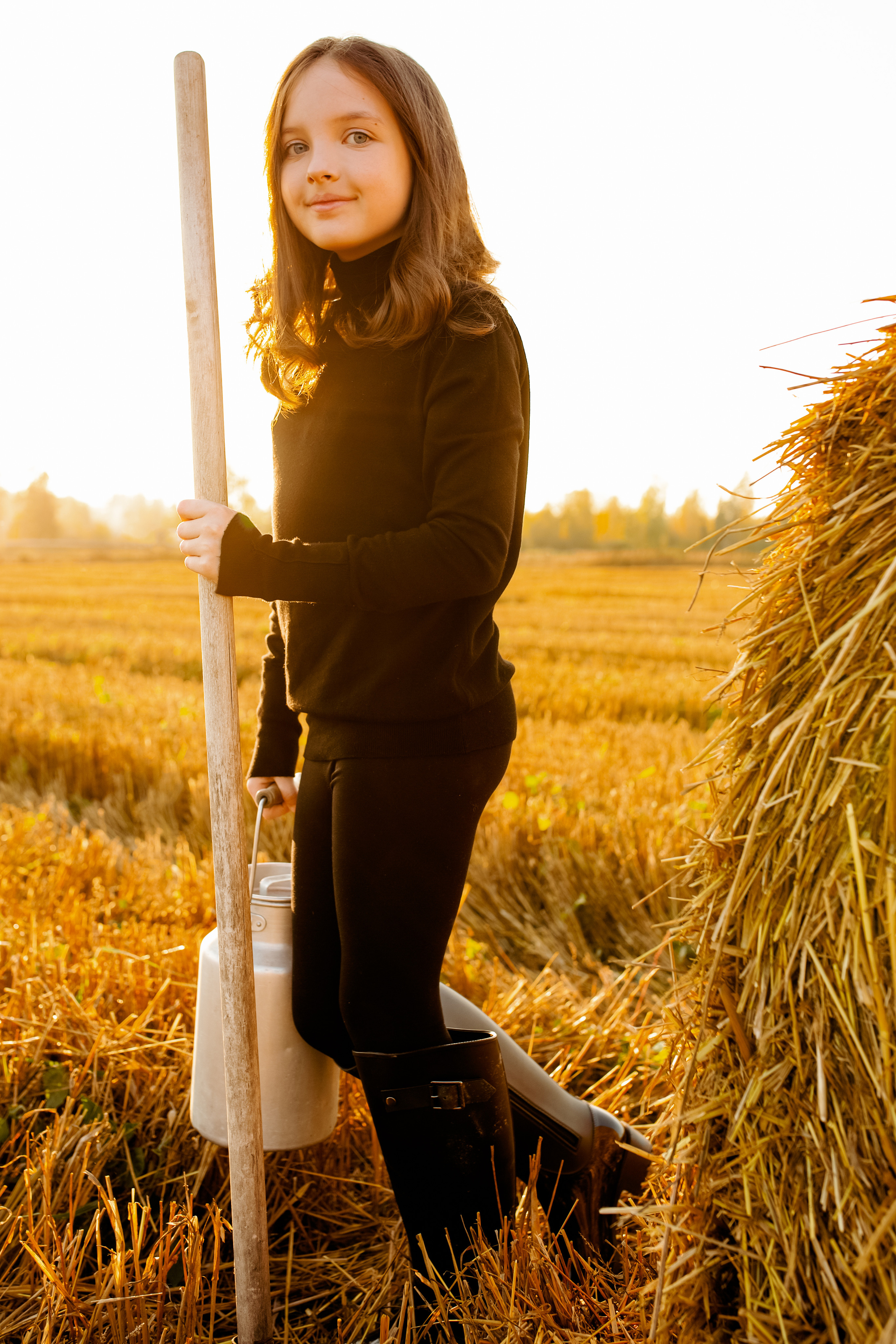 Девочка в черном с граблями и бидоном молока в поле на закате.
