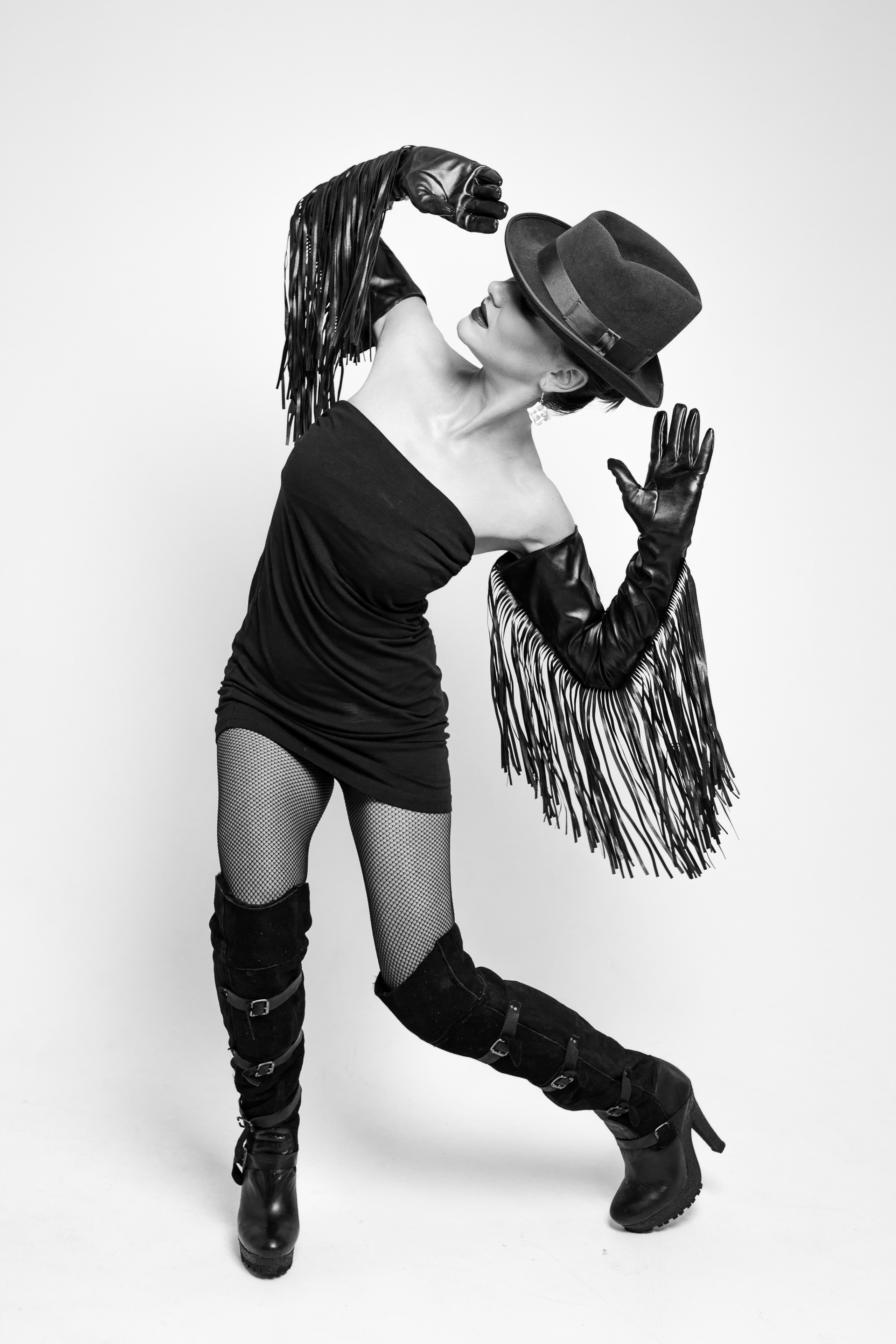 Черно-белая фотография девушки в шляпе, длинных перчатках с бахромой и ботфортах, которая танцует.