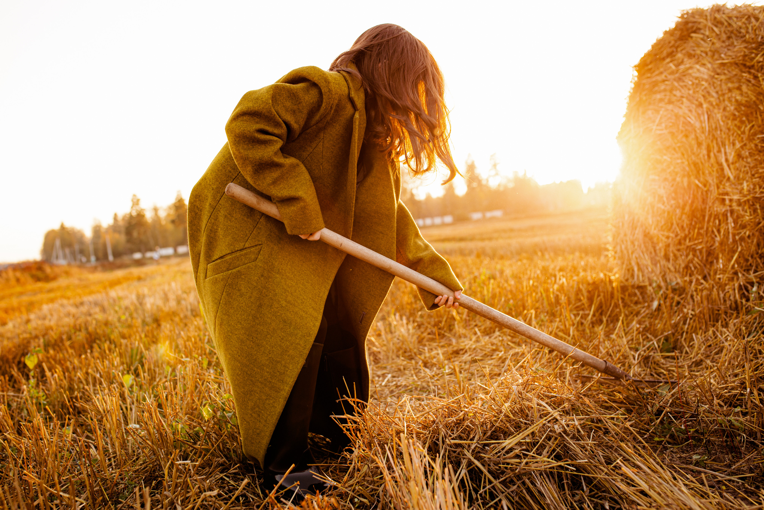 Детская осенняя фотосессия в поле на закате со стогами сена. Девочка в пальто работает граблями в пшеничном поле.