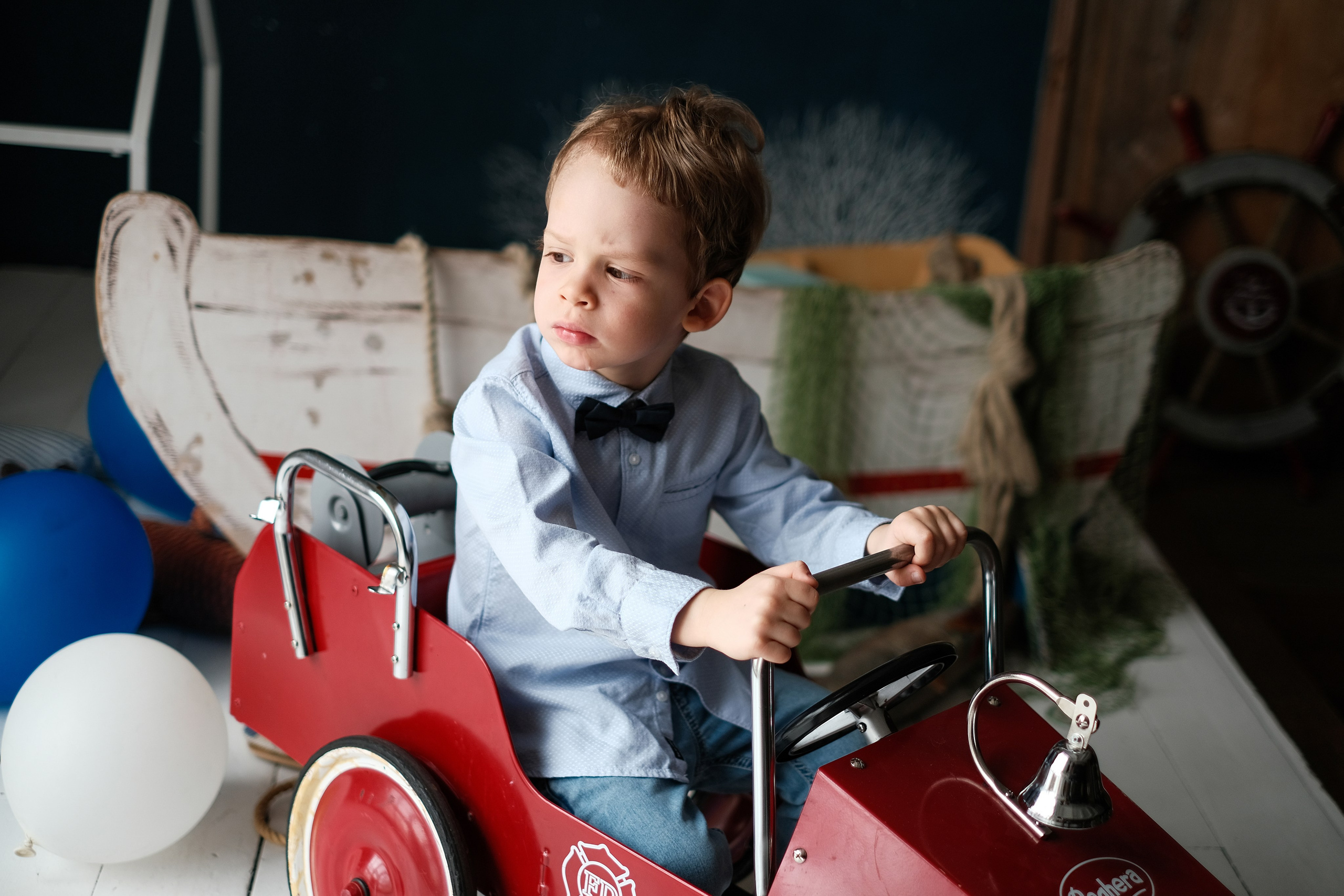 Мальчик в галстуке-бабочка сидит в красной игрушечной машине.