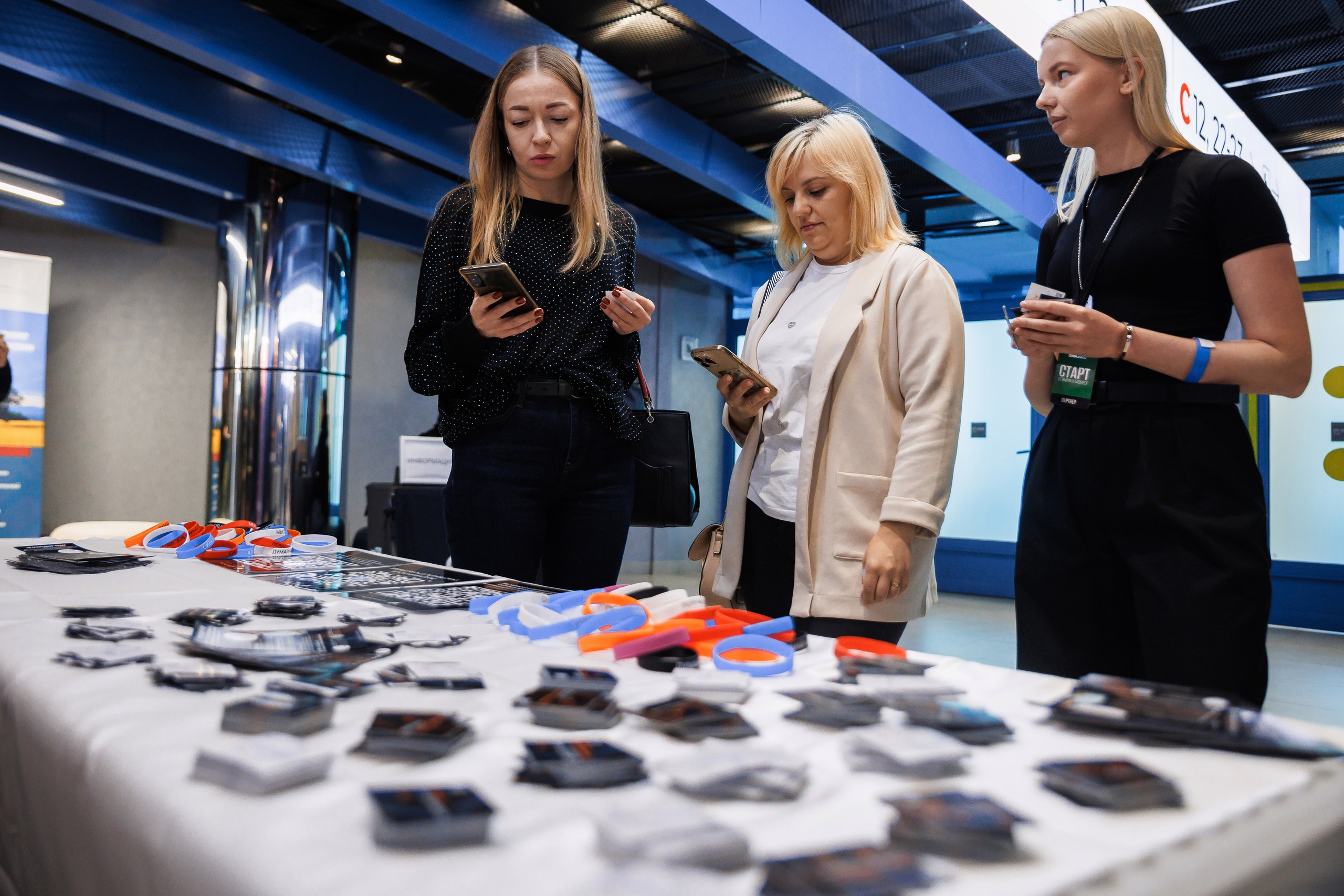 Три женщины стоят у стола, заваленного карточками, листовками и оранжево-синими браслетами, на мероприятии в помещении. Две смотрят в свои телефоны, а одна — на стол. Обстановка выглядит современной и профессиональной.