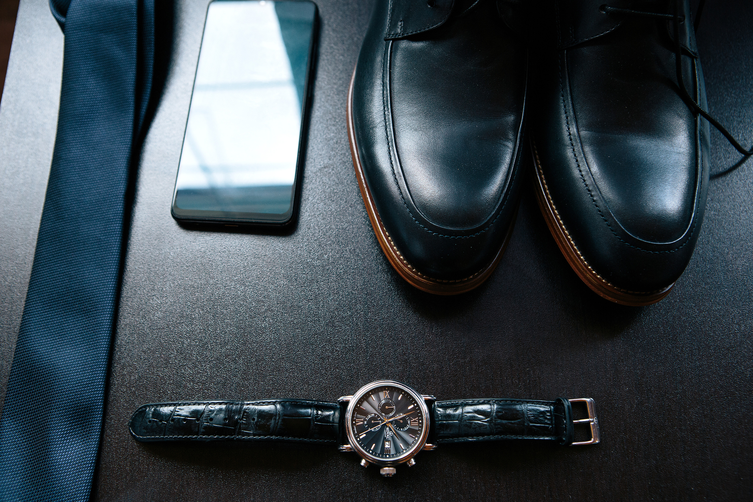 Туфли, часы, галстук и телефон жениха на темном столе