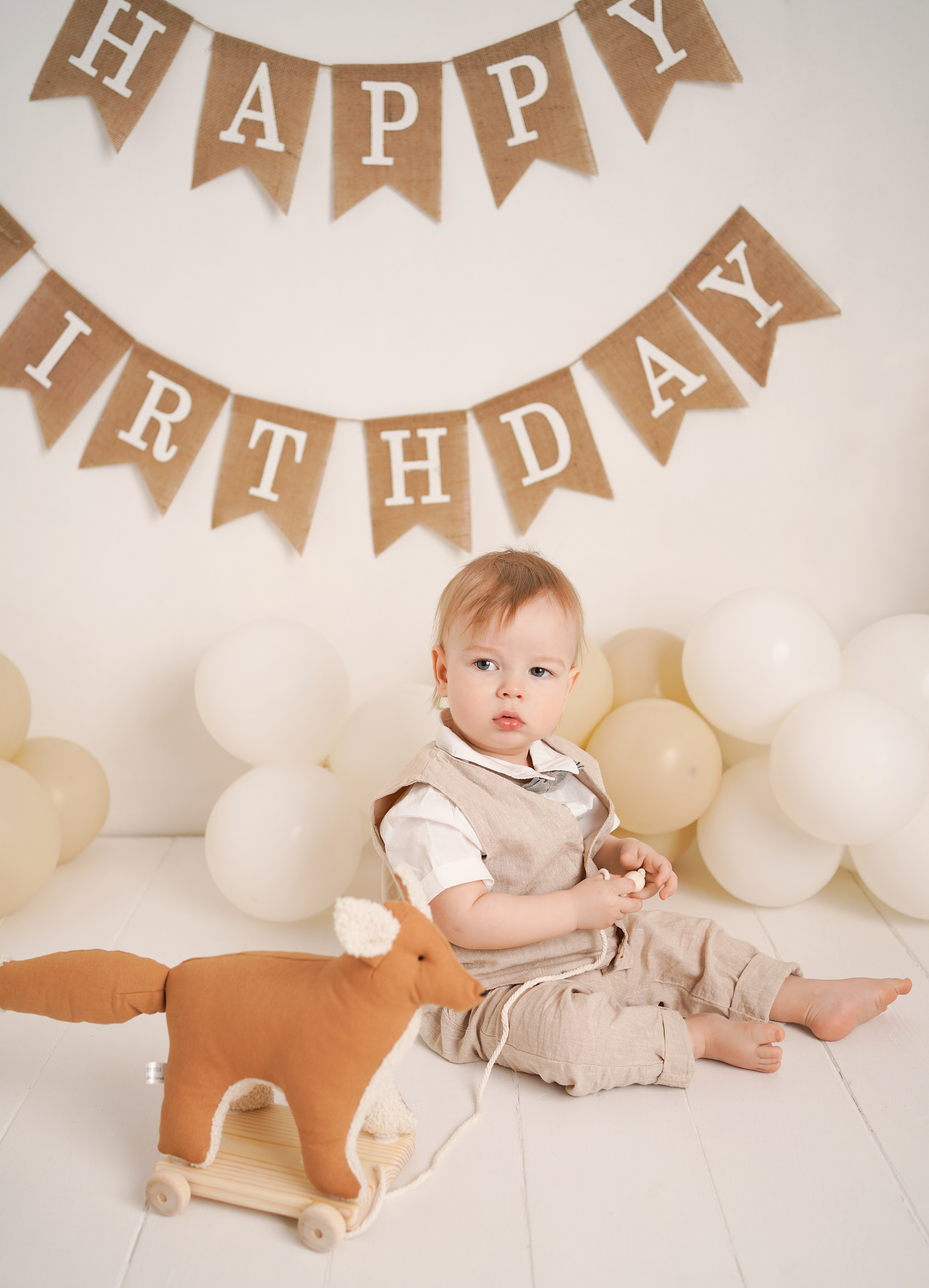 На фото малыш с игрушками ,  бежевыми шариками и гирляндой С днем рождения.