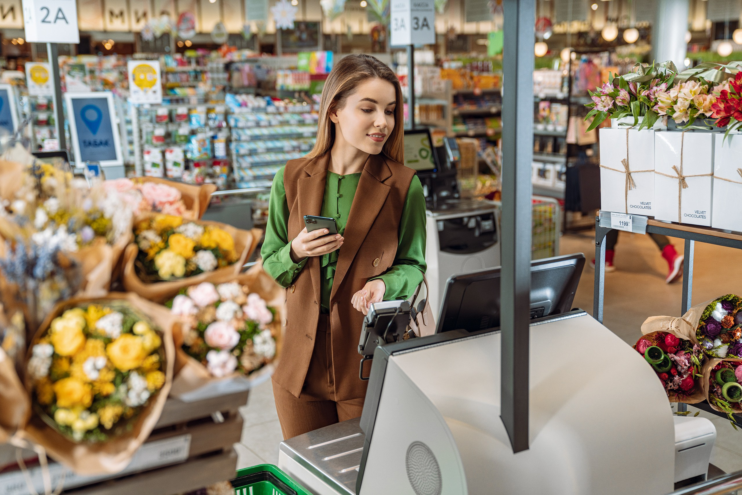 Девушка в окружении цветов на кассе самообслуживания оплачивает товар 