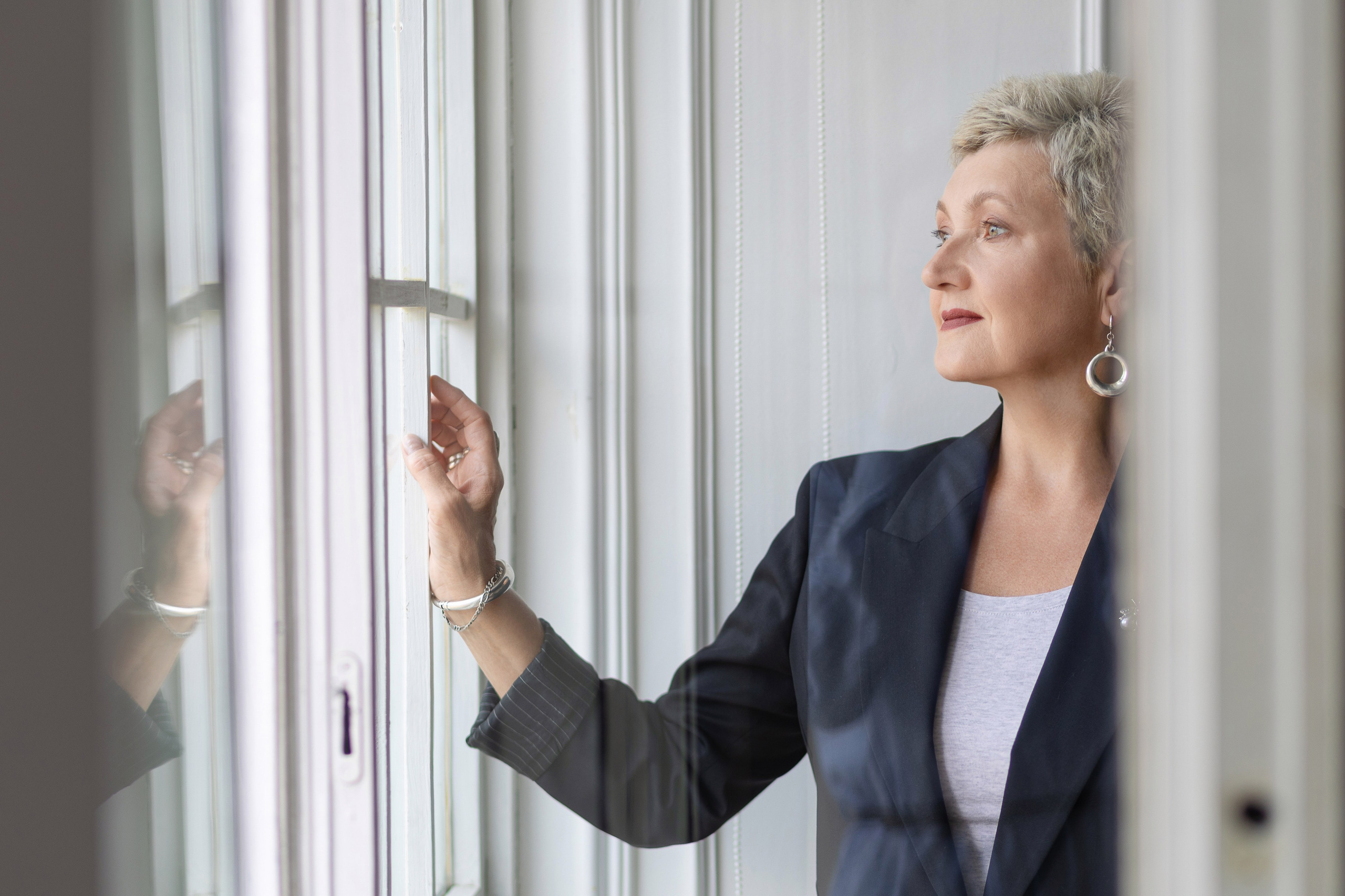 Женщина стоит у окна, правой рукой держась за створку.
