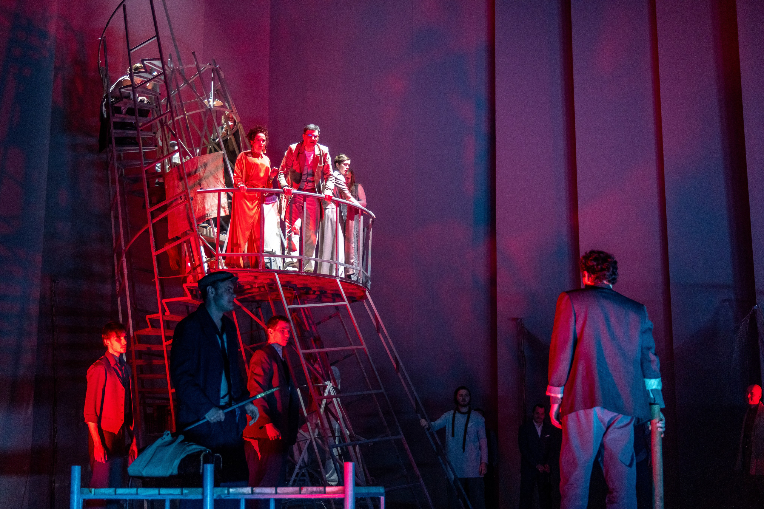 Группа актёров в красных и серых костюмах стоит на металлической лестнице, освещённые красным светом.