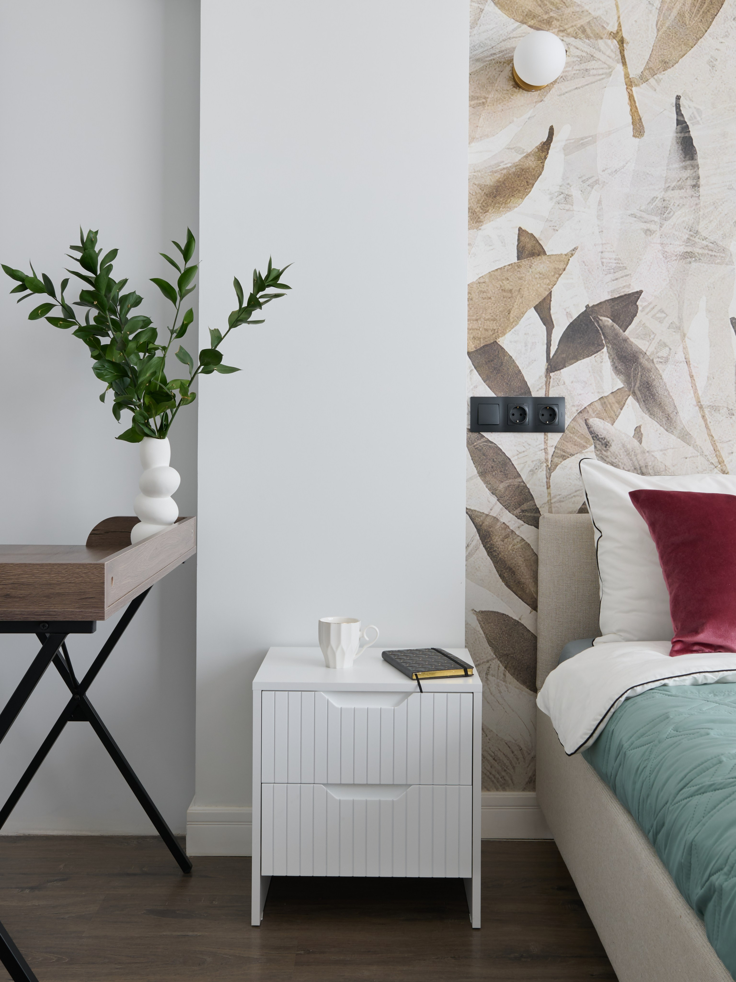 Уголок спальни с рабочим столом и белым комодом. На стене висит абстрактное изображение, а на столе и комоде расставлены декоративные элементы и зеленые растения.