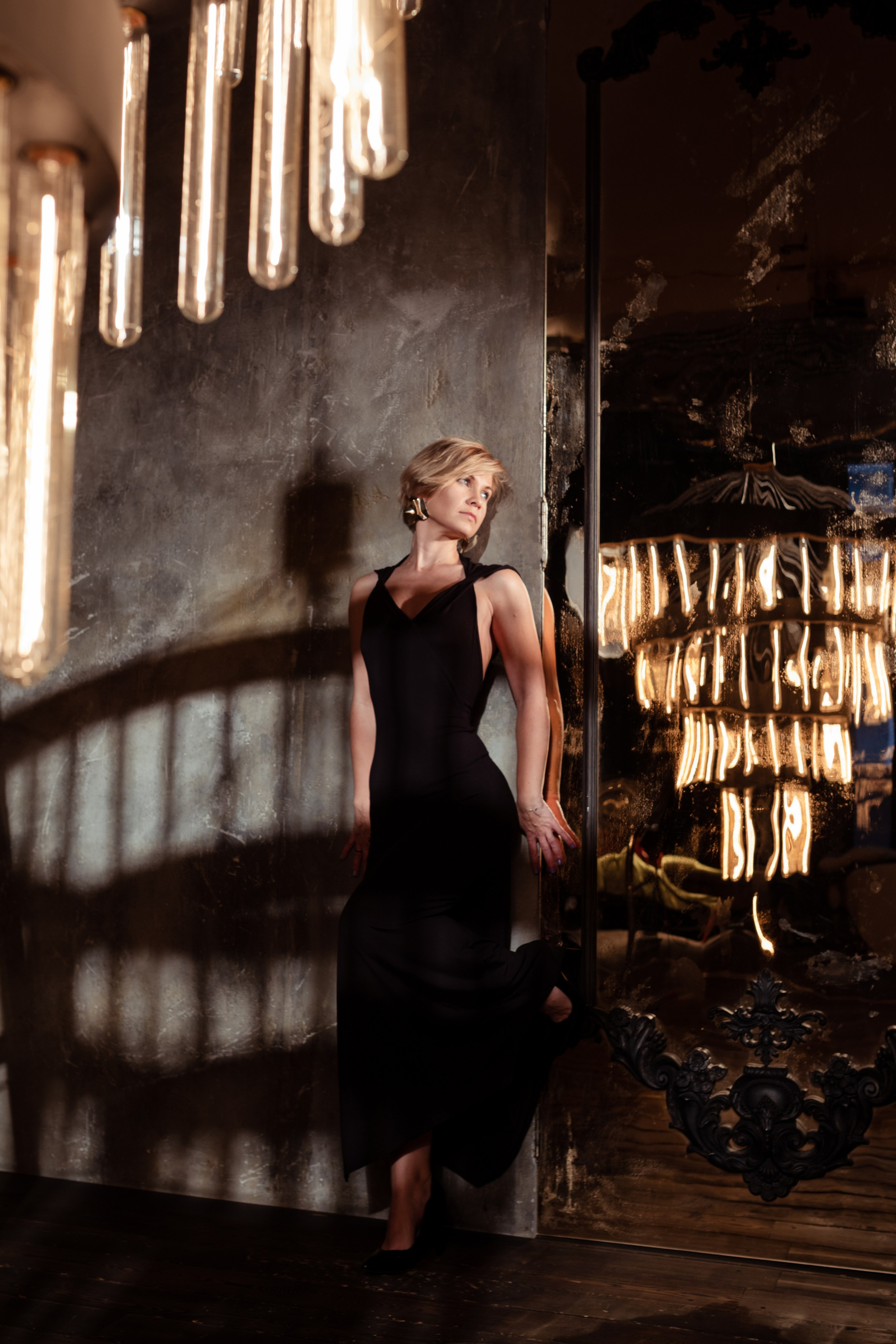 Женщина в чёрном платье стоит у стены в зале с огромной люстрой