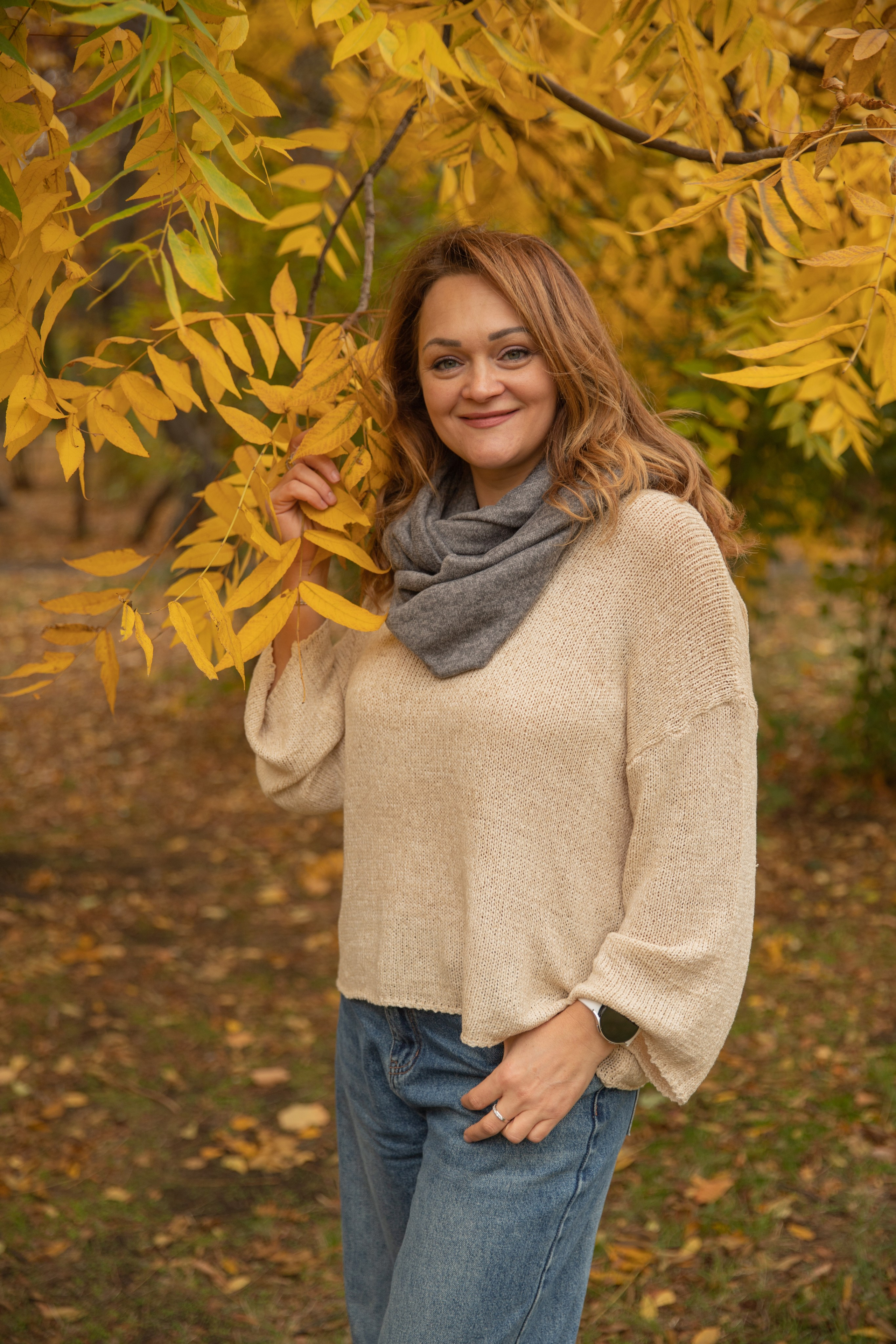 Красавица в уютном свитере прогуливается по осеннему парку Чистяковская роща, окружённая желтыми листьями и теплым светом.