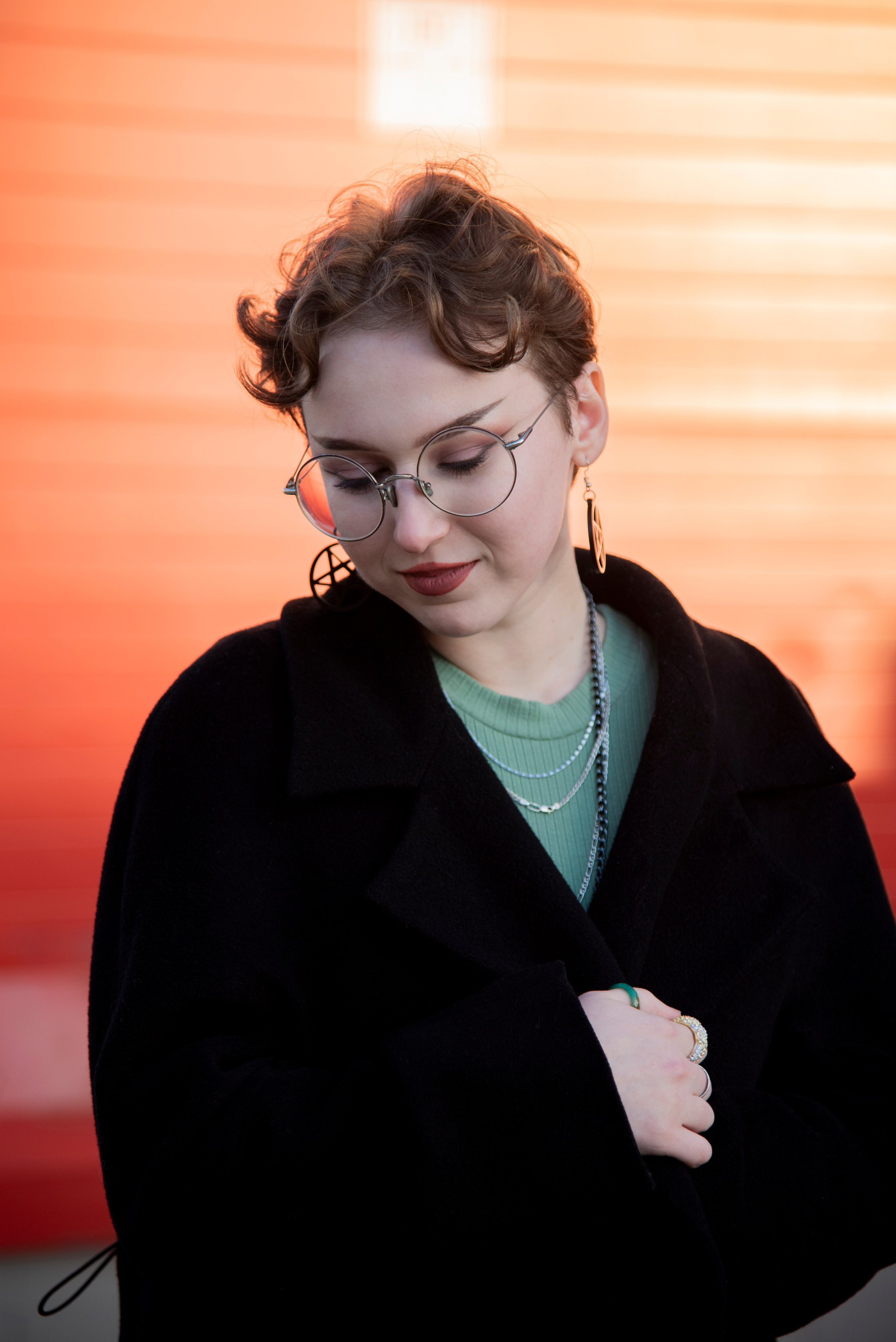 портет девушки. чёрное пальто. красивые позы. серьки в виде пентограммы. короткая стрижка. круглые очки. красный фон