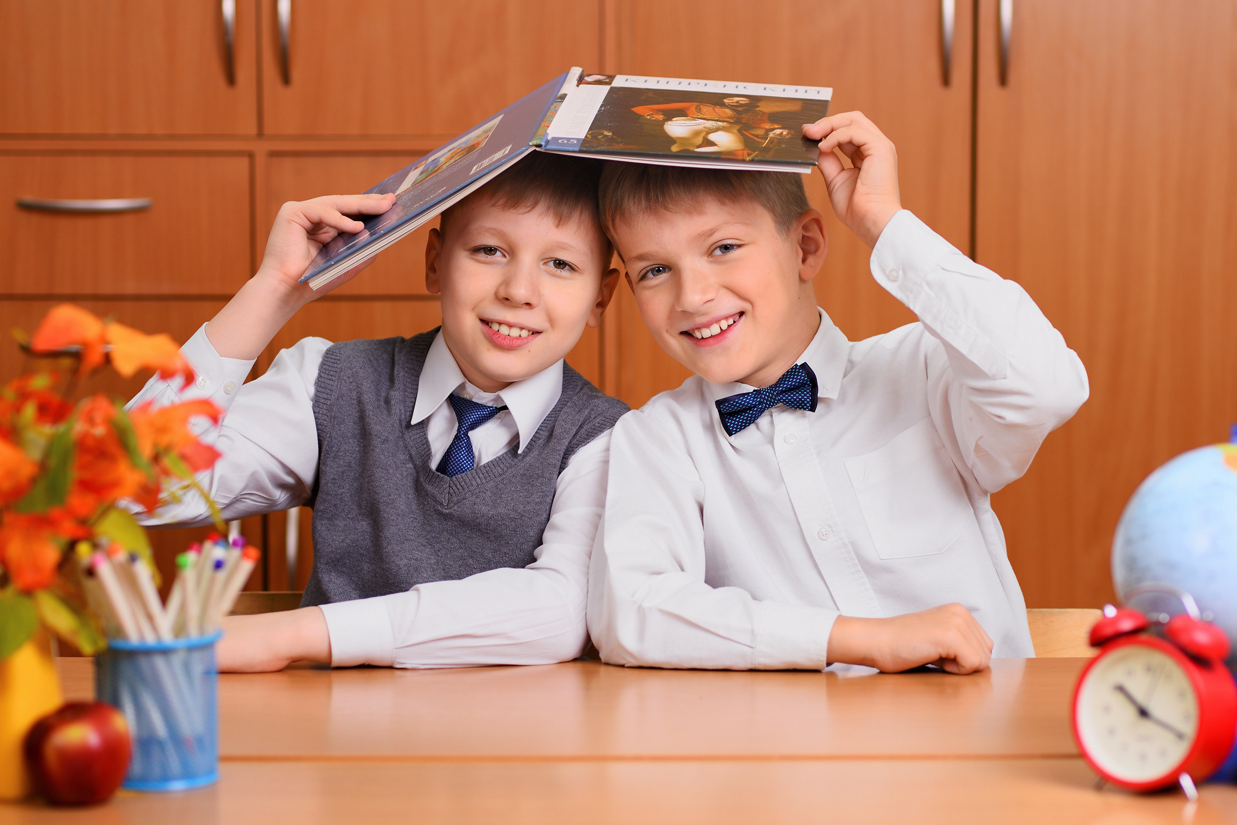Улыбающиеся школьники сидят за партой и держат над головой книгу, как крышу домика.