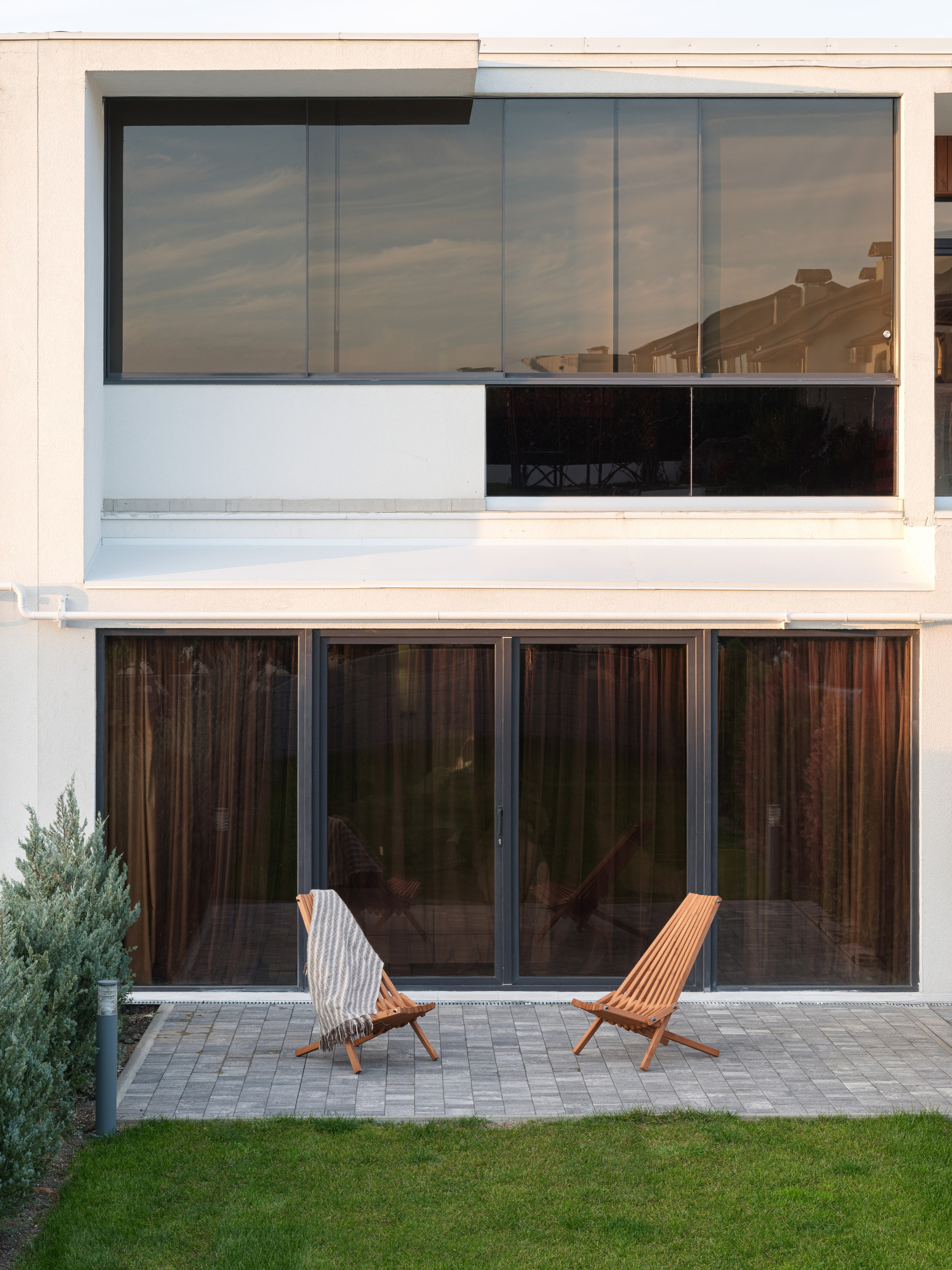 Фасад современного дома с панорамными окнами и уютной террасой, оформленной деревянными шезлонгами и мягким пледом на фоне ухоженного газона. Архитектурная композиция подчеркивает минимализм и уют проекта дизайнера Алины Дёровой, стилист Наталья Якушева, фото Сергей Денисенко.