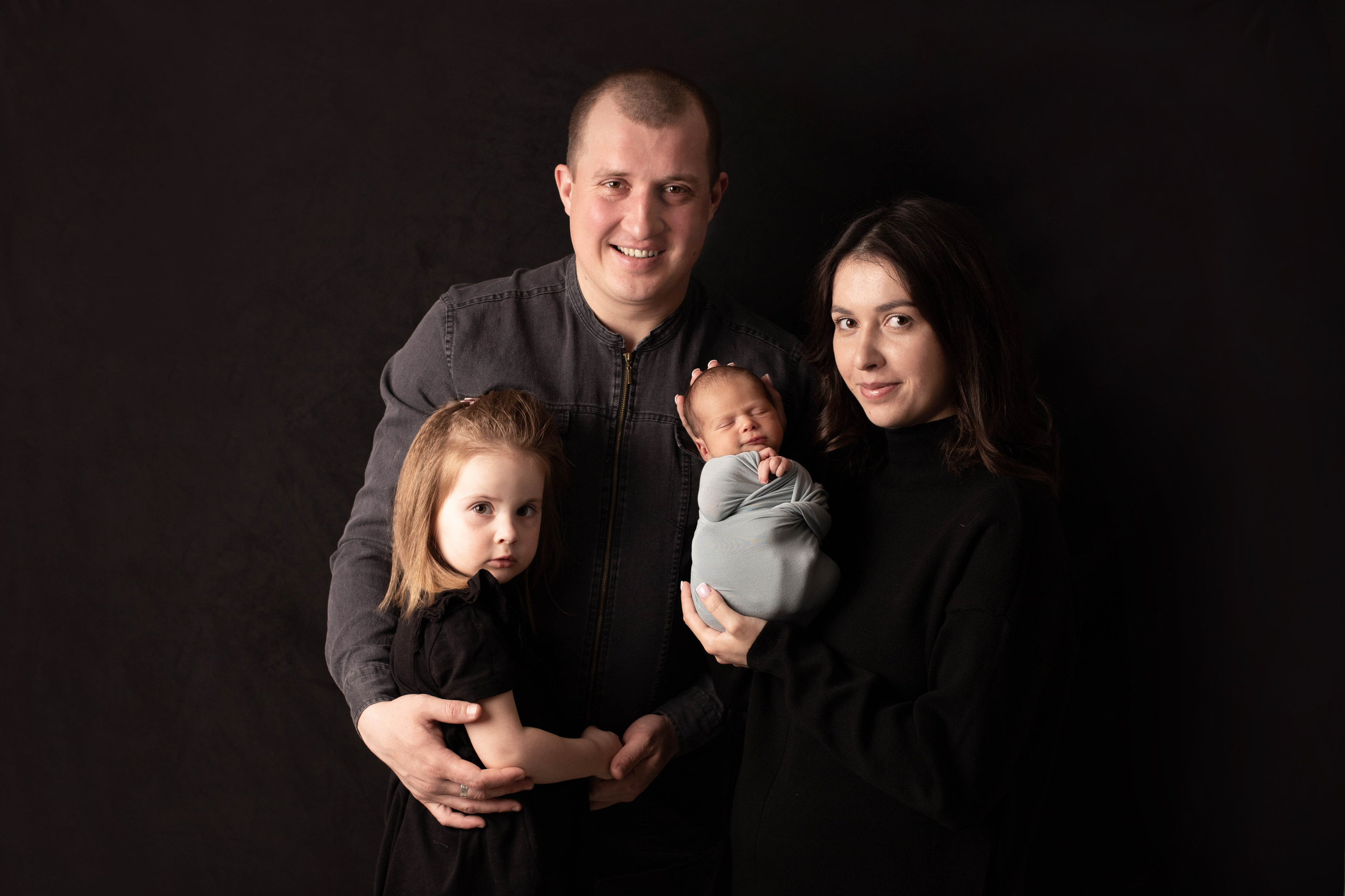 Фотография  счастливой семьи с новорожденным и старшей дочкой на темном фоне. Мама держит малыша на руках.