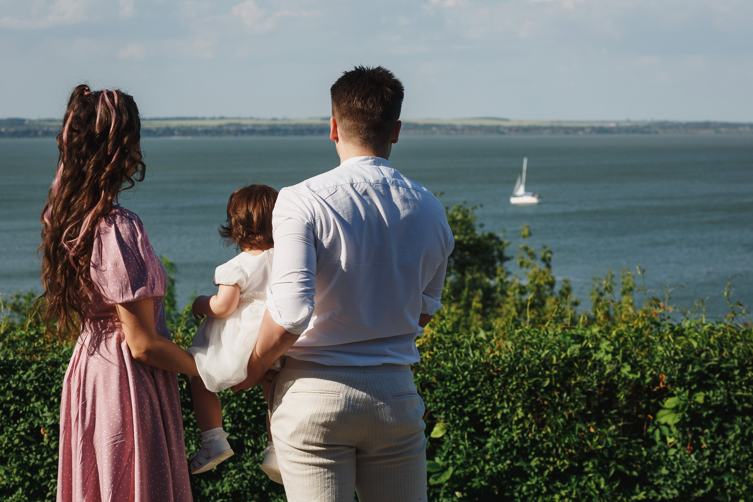 Семейная съёмка в Таганроге: родители с маленьким ребенком на руках смотрят на яхту в Таганрогском заливе. Фотография сделана на фоне живописного вида из ресторана Feola's Kitchen & Wine.