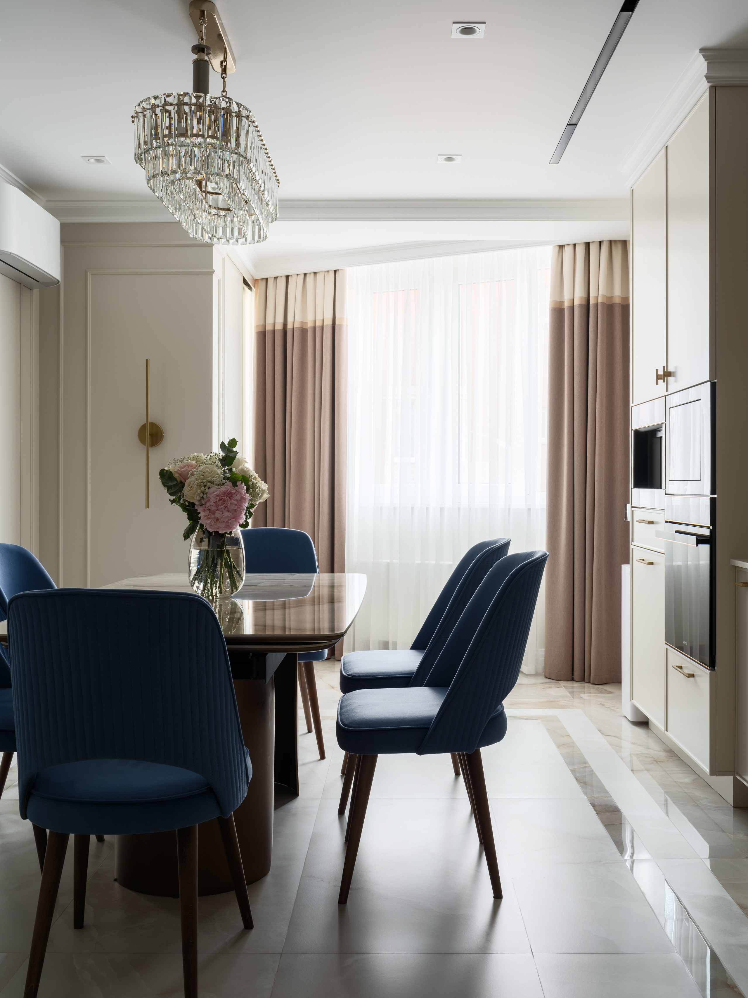 Элегантная столовая зона на кухне с прямоугольным обеденным столом с глянцевой столешницей и синими мягкими стульями на деревянных ножках, украшенная хрустальной люстрой и дополненная двойными шторами, дизайн интерьера Евгении Ермошиной, фото Сергея Денисенко.