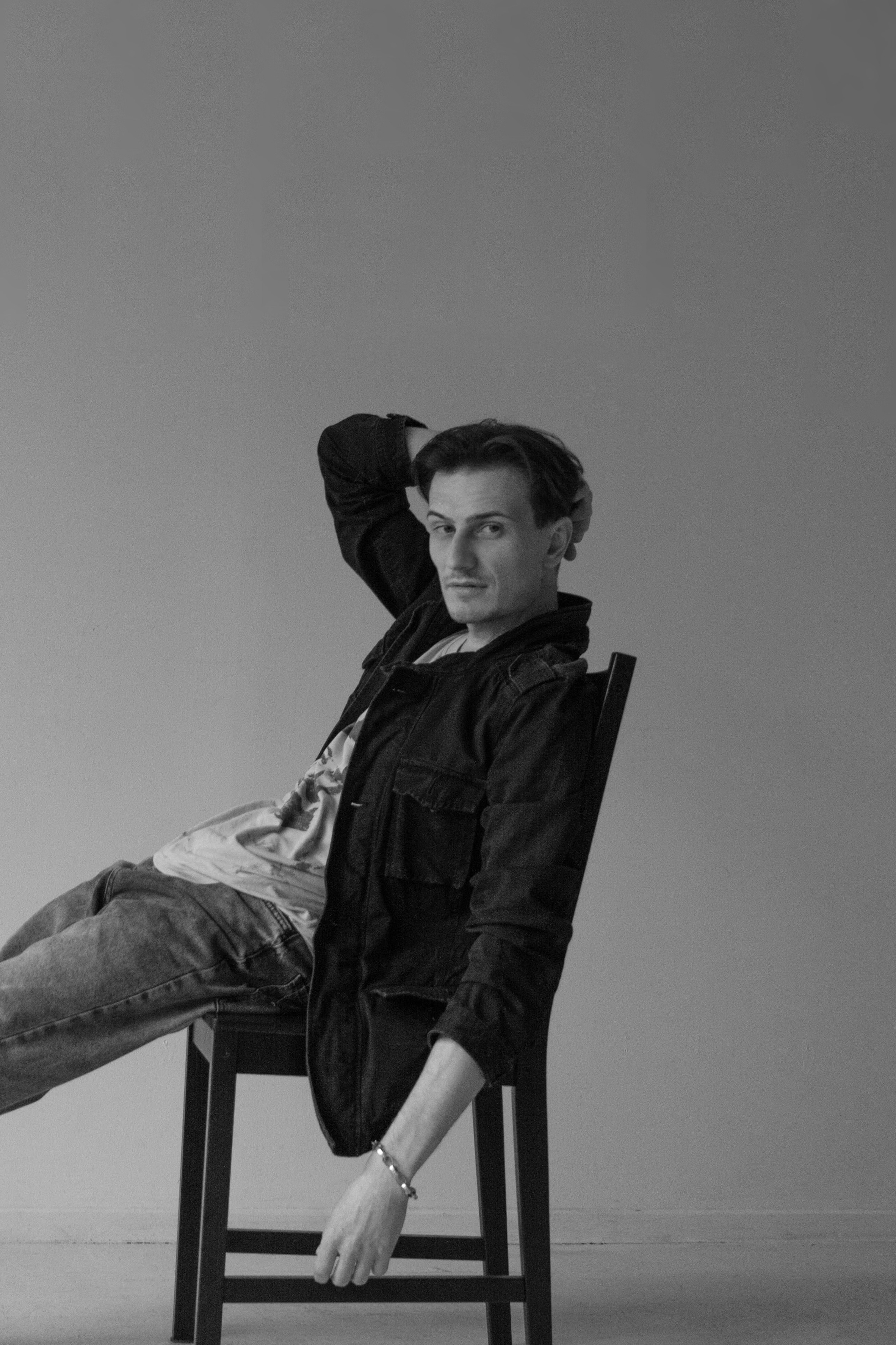 Мужчина сидит на стуле, закинув руку за спину на сером фоне.