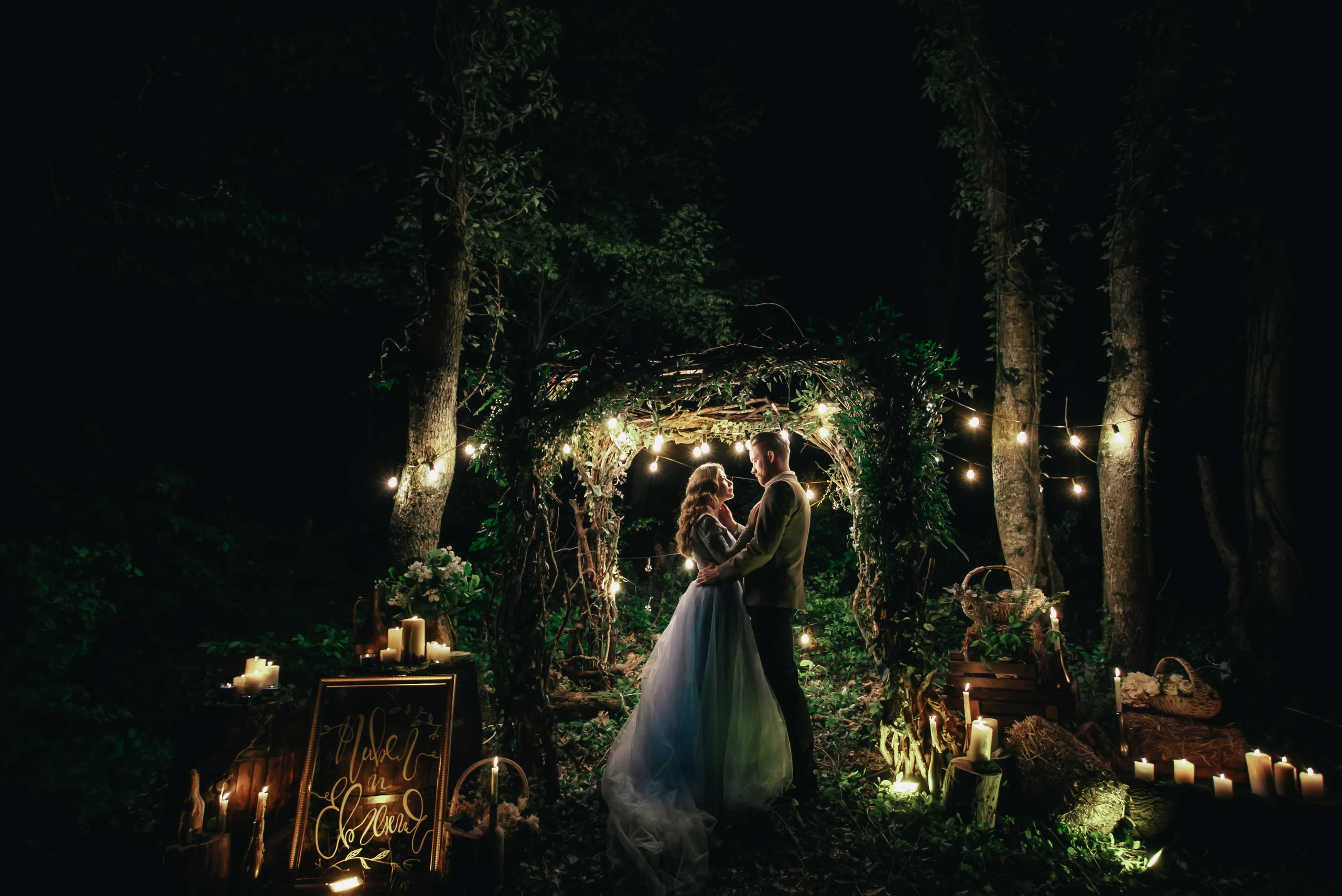 Жених и невеста в голубом свадебном платье с каштановыми распущенными волосами стоят взявшись за руки ночью в лесу украшенном гирляндами