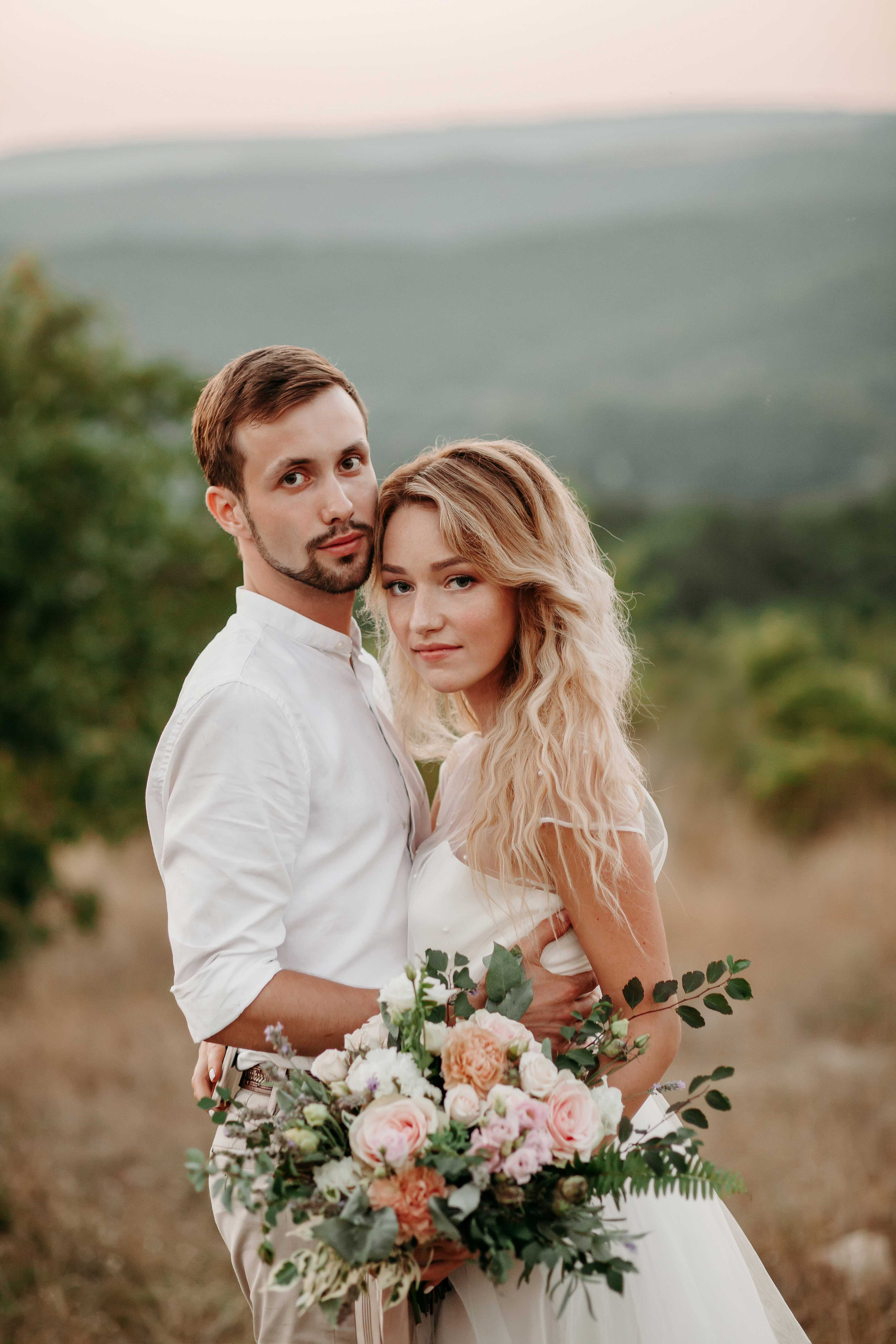 Невеста со светлыми распущенными волосами в белом платье стоит в обнимку с  женихом  в белой рубашке