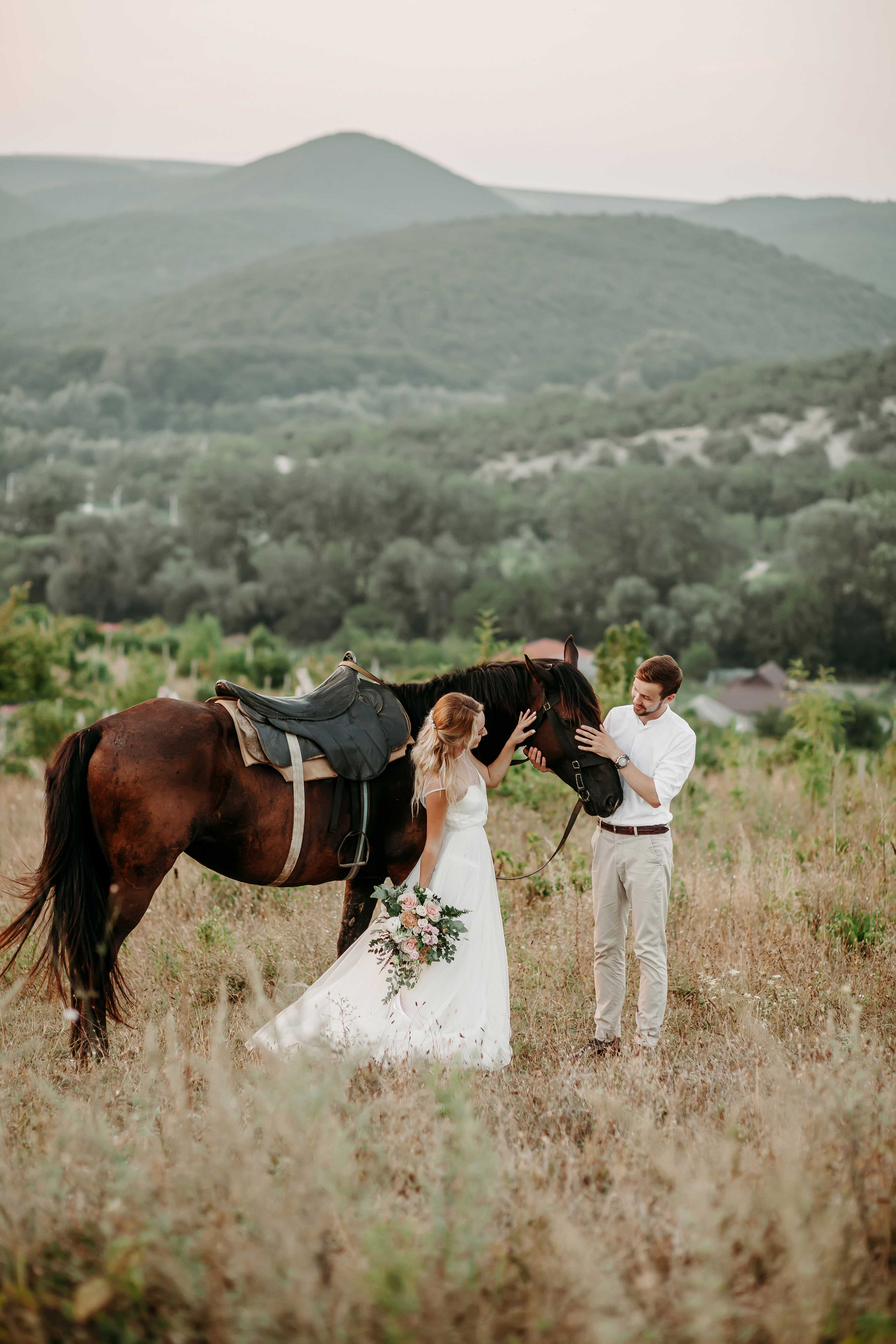 Блондинка невеста в белом платье и жених в бежевых брюках стоят рядом с лошадью