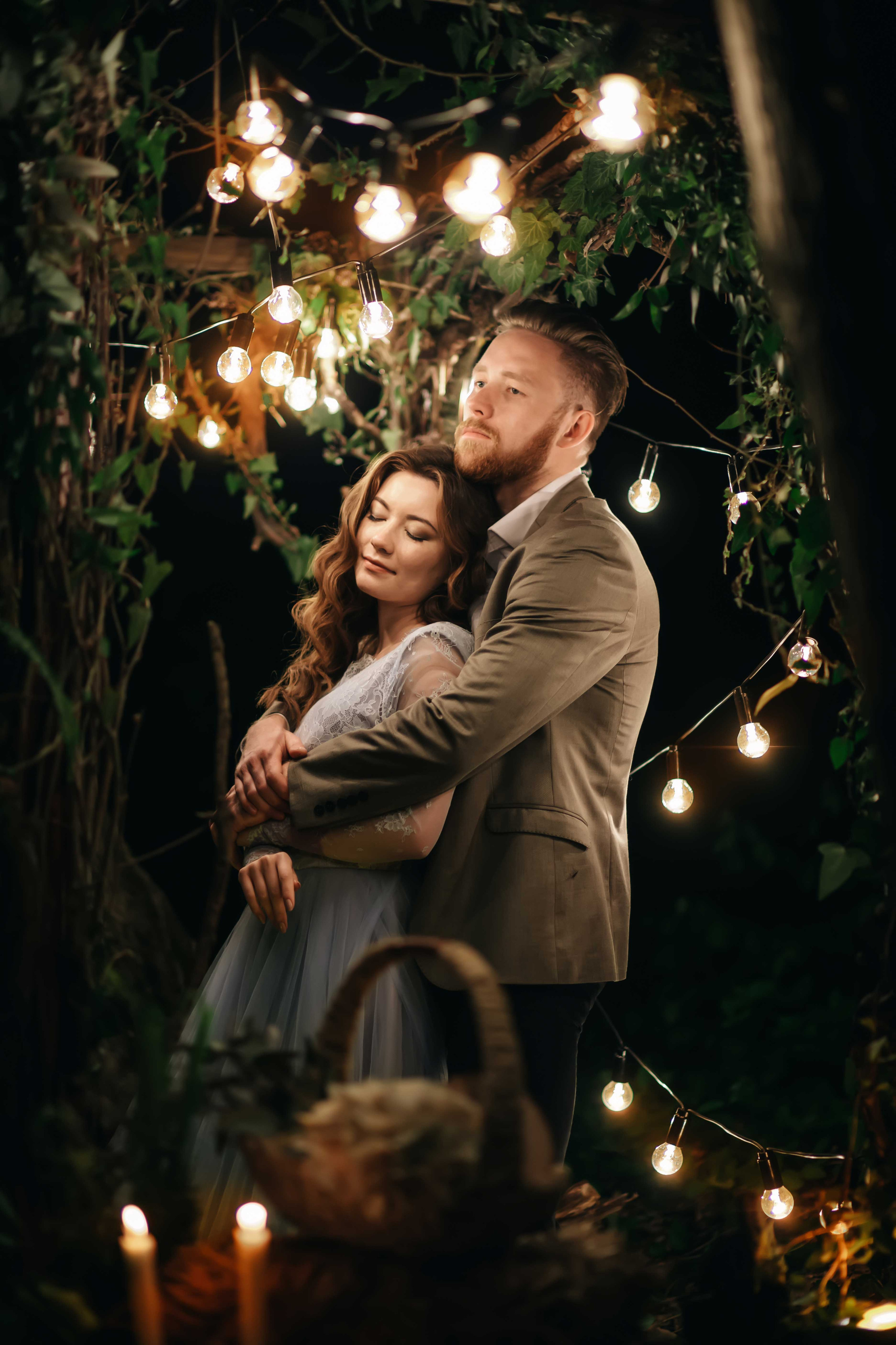 Жених обнимает невест в голубом свадебном платье с каштановыми распущенными волосами ночью в лесу украшенном гирляндами