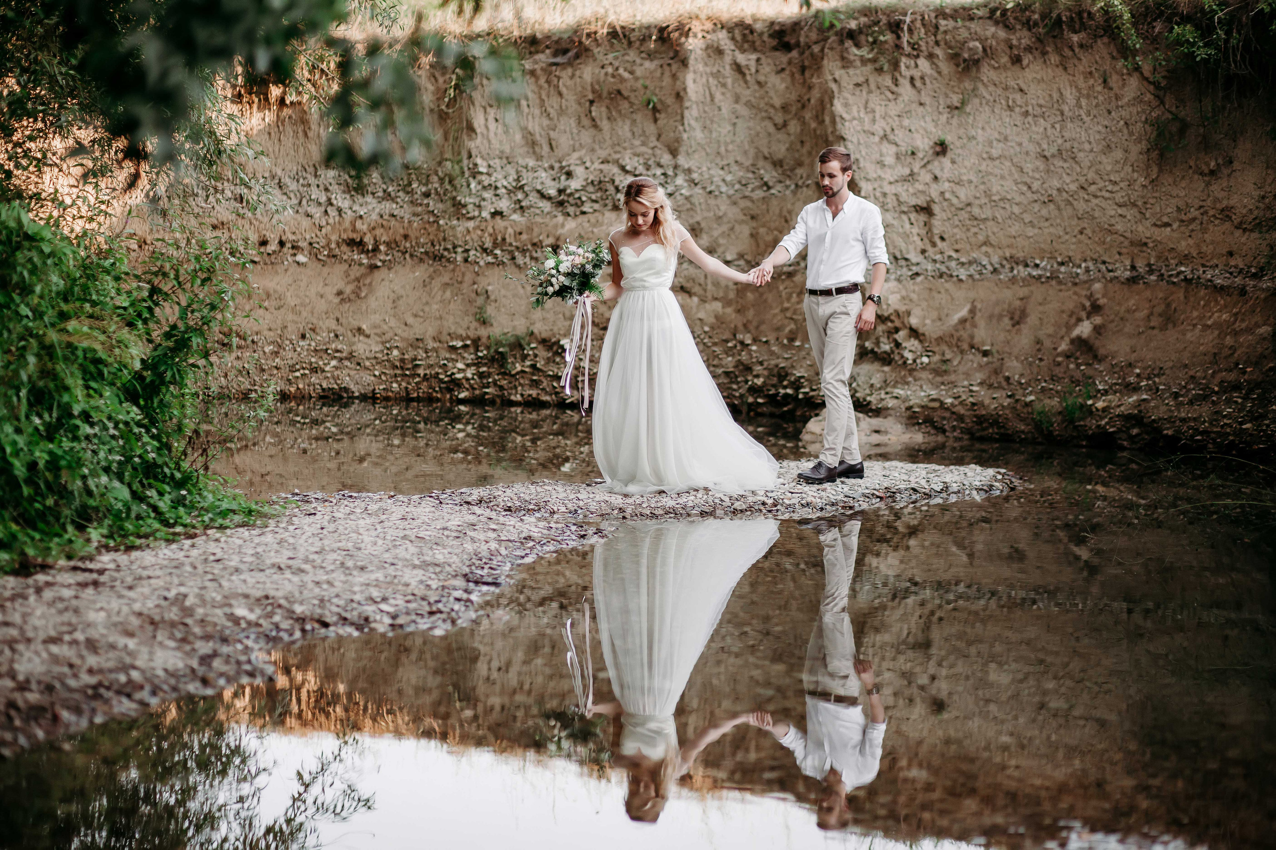 Невеста с распущенными светлыми волосами и жених в белой рубашке идут по берегу реки взявшись за руки