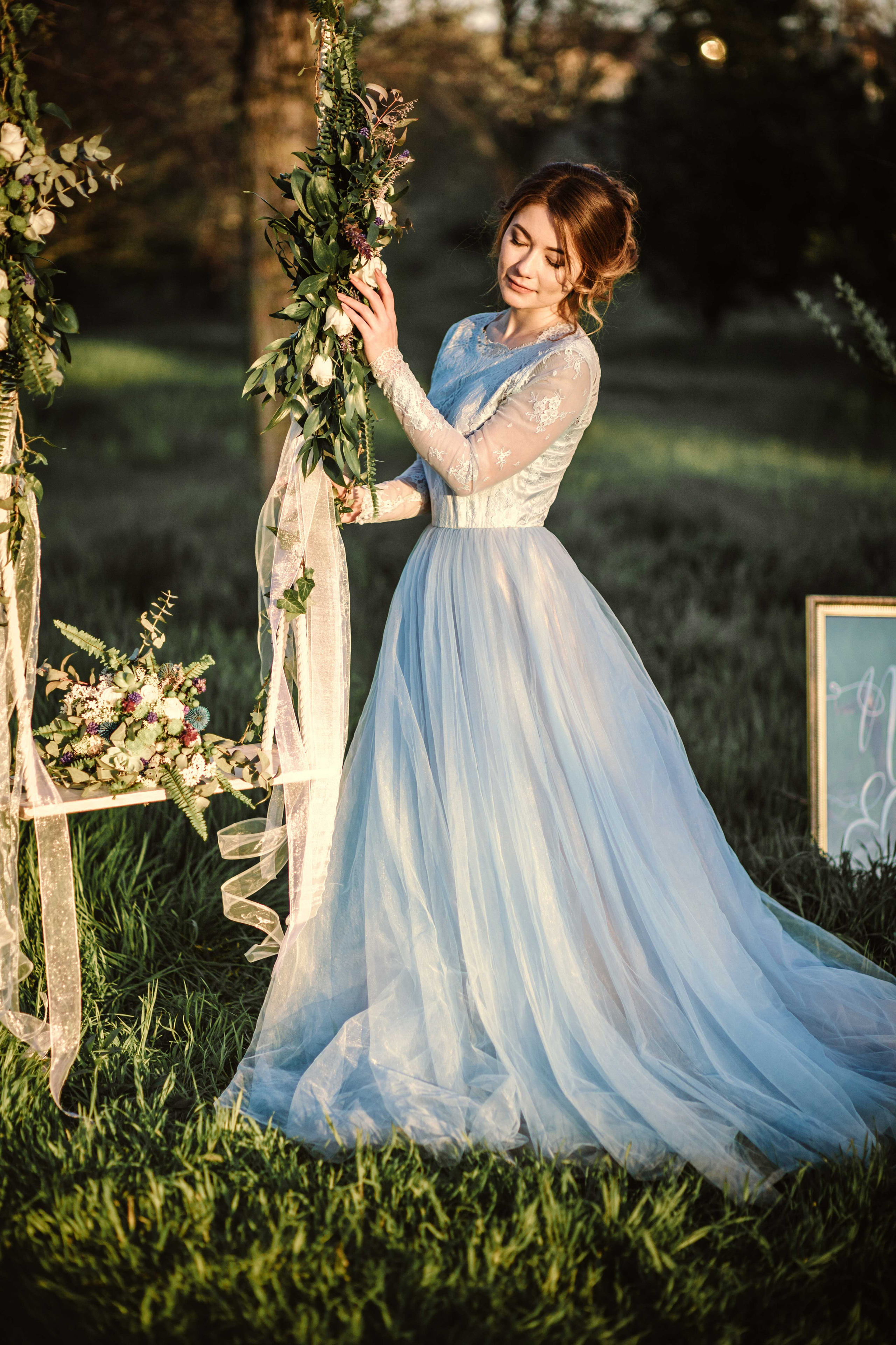 Красивая невеста в голубом свадебном платье стоит рялом с качелями украшенными цветами