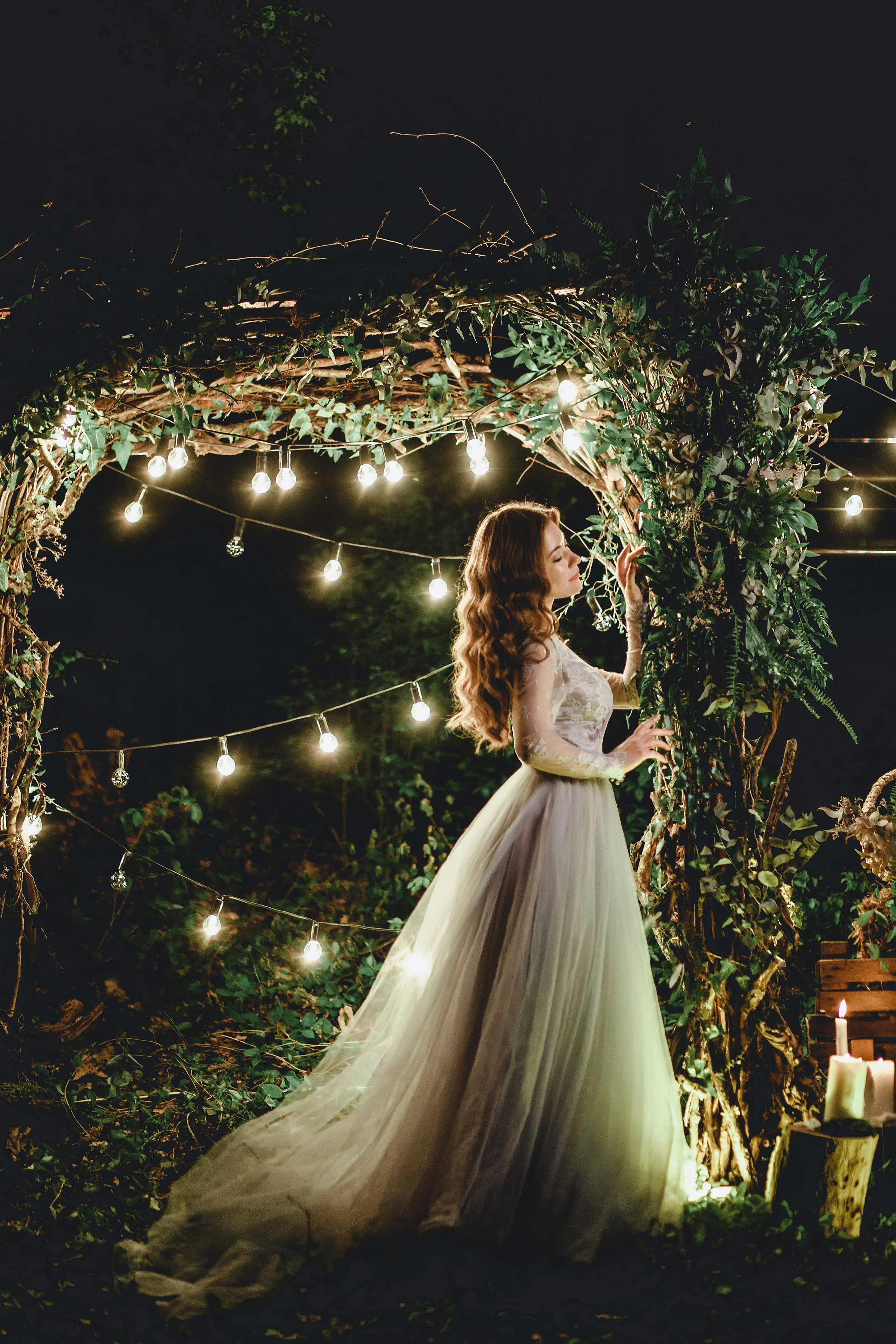 Невеста в голубом свадебном платье с каштановыми распущенными волосами ночью в лесу украшенном гирляндами