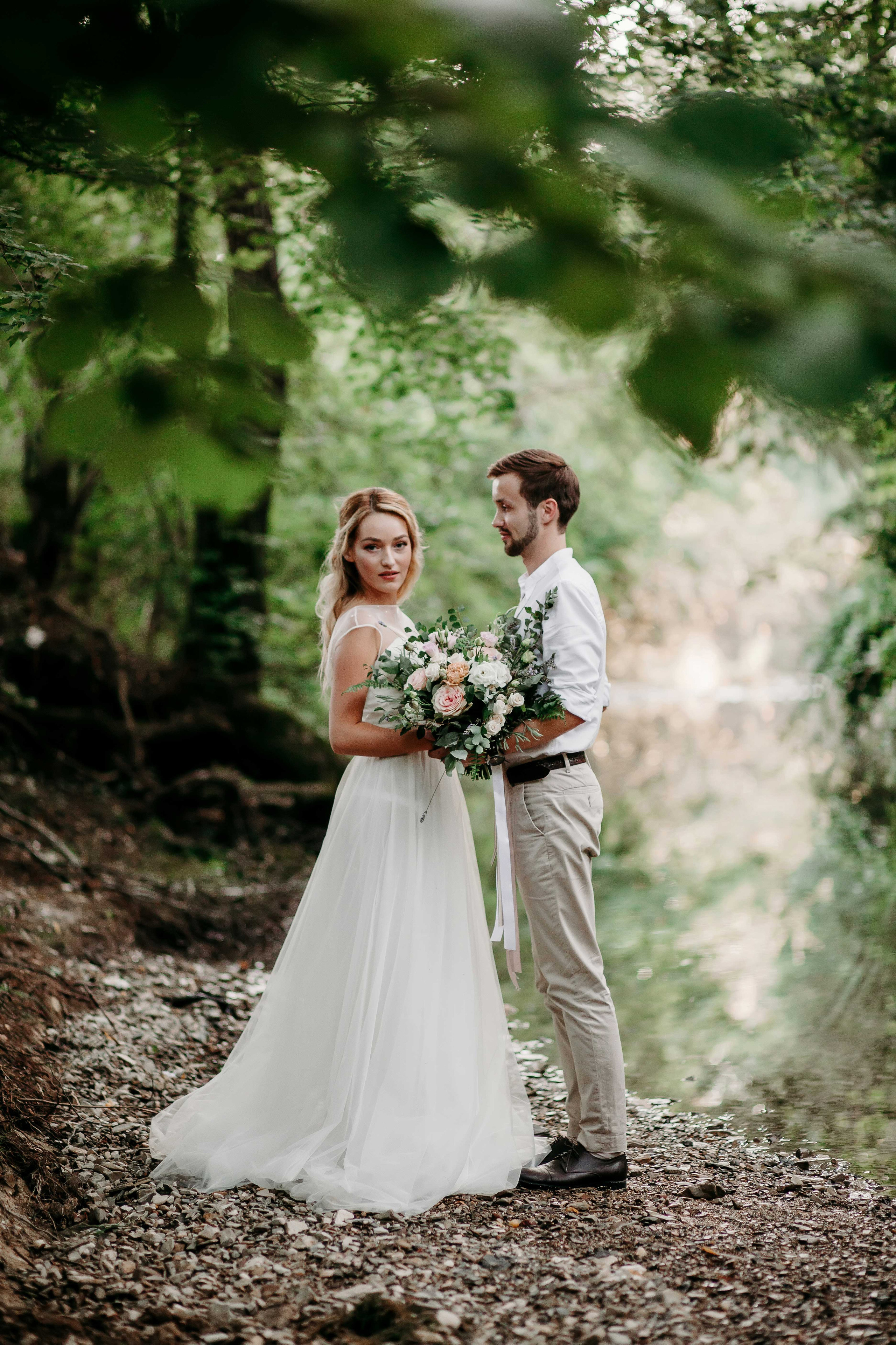 Невеста с распущенными светлыми волосами и жених в белой рубашке стоят у реки взявшись за руки