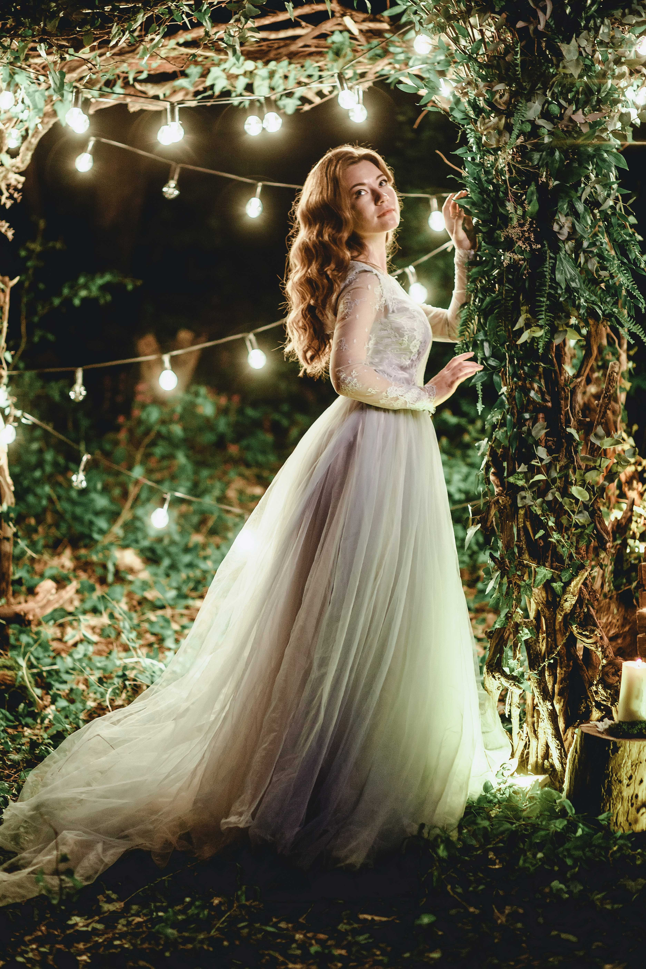 Невеста в голубом свадебном платье с каштановыми распущенными волосами ночью в лесу украшенном гирляндами