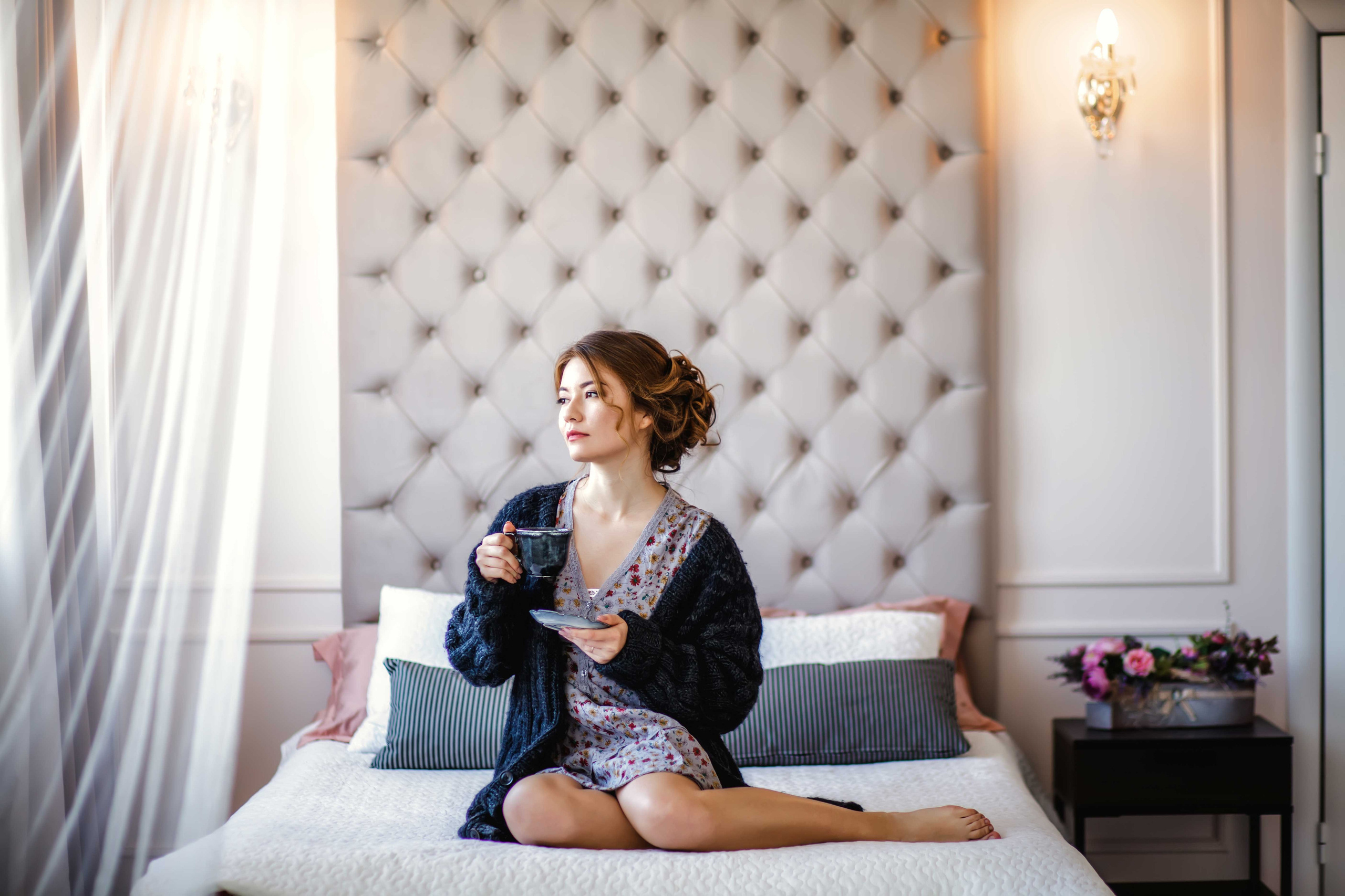 Девушка с каштановыми волосами сидит на кровати с чашкой кофе и смотрит в окно
