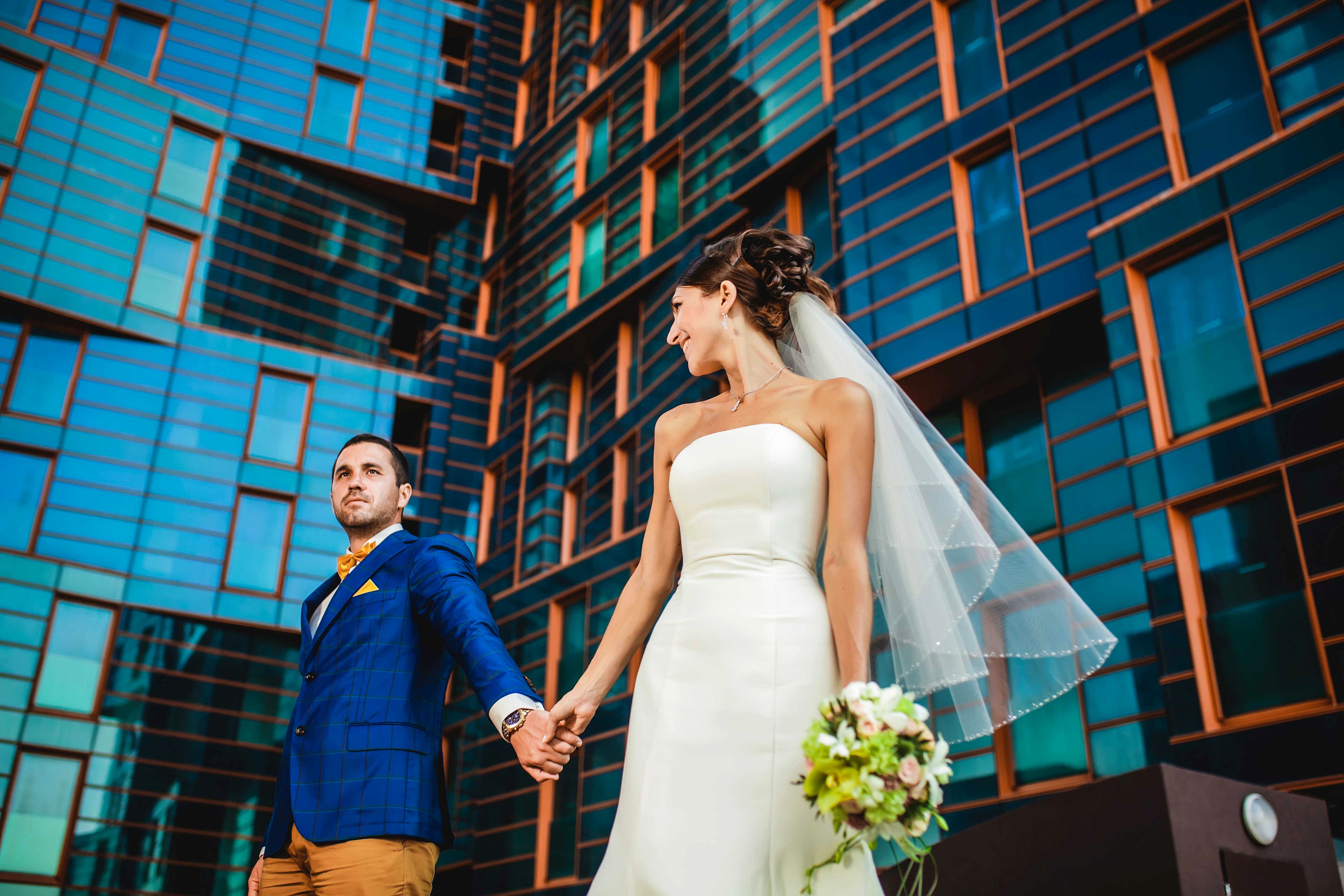 Жених и невеста стоят взявшись за куки на фоне высокого красивого здания