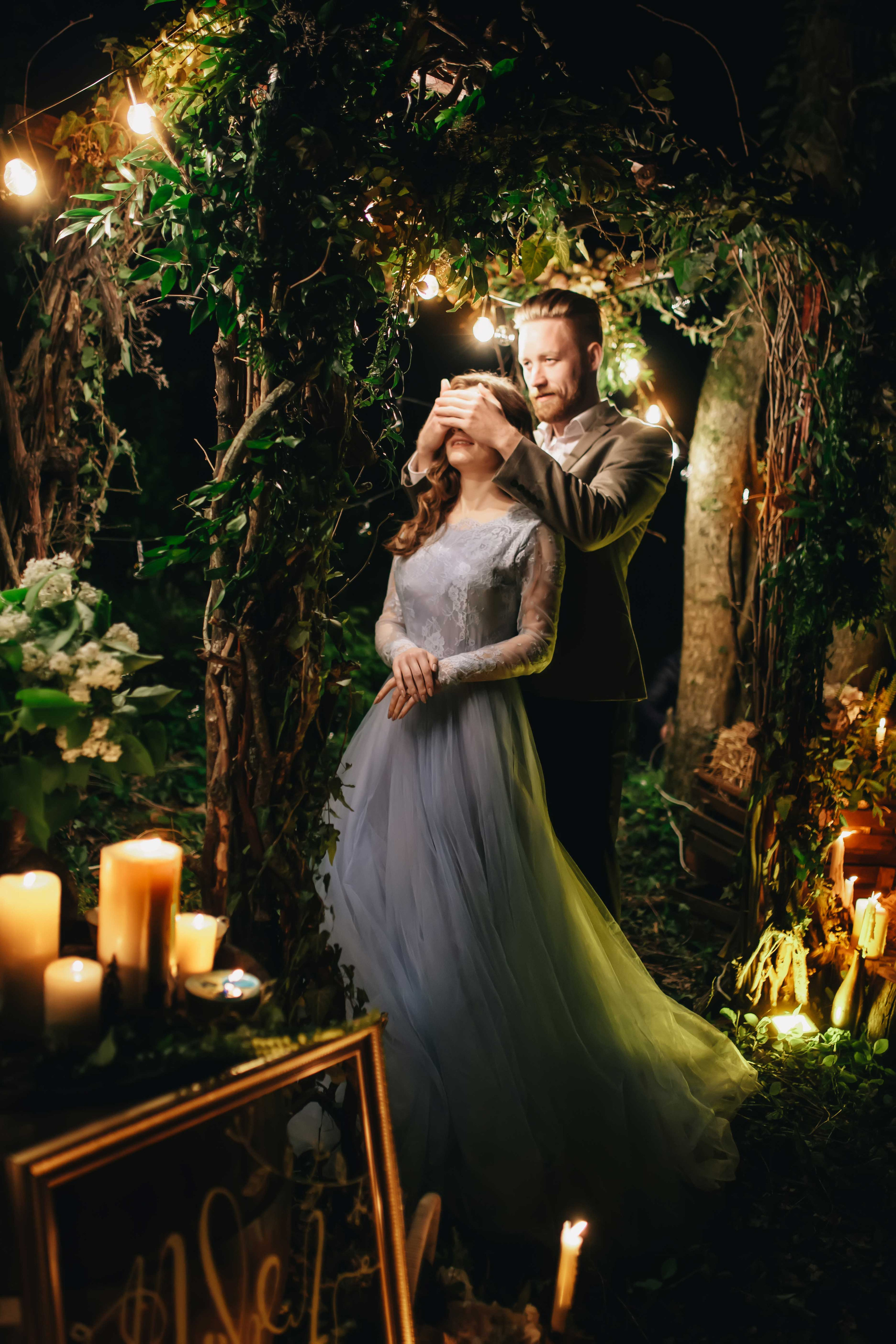 Жених закрывает глаза невесте в голубом свадебном платье с каштановыми распущенными волосами ночью в лесу украшенном гирляндами