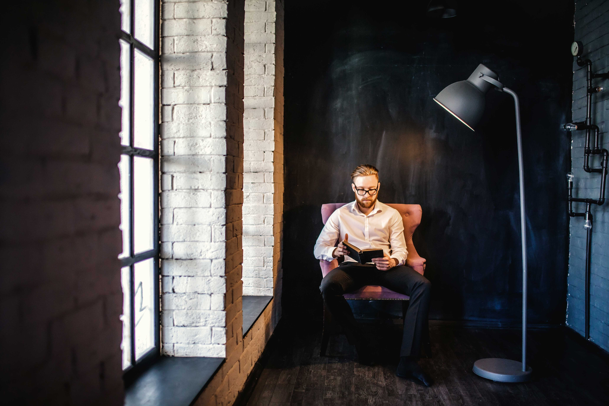 Молодой человек в очках читает книгу сидя в кресле