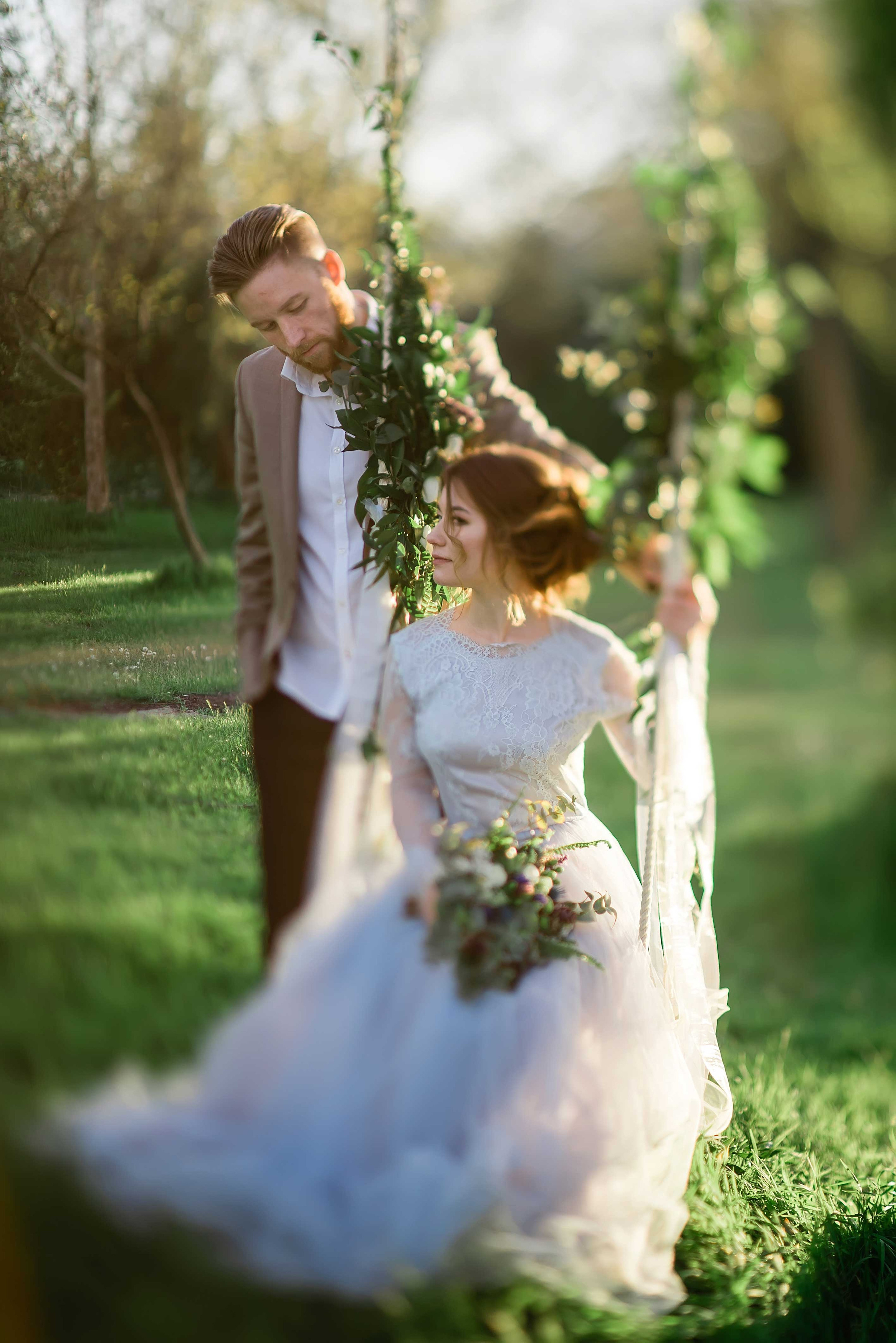 Нежная стильная невеста в голубом свадебном платье сидит на качелях а рядом стоит жених