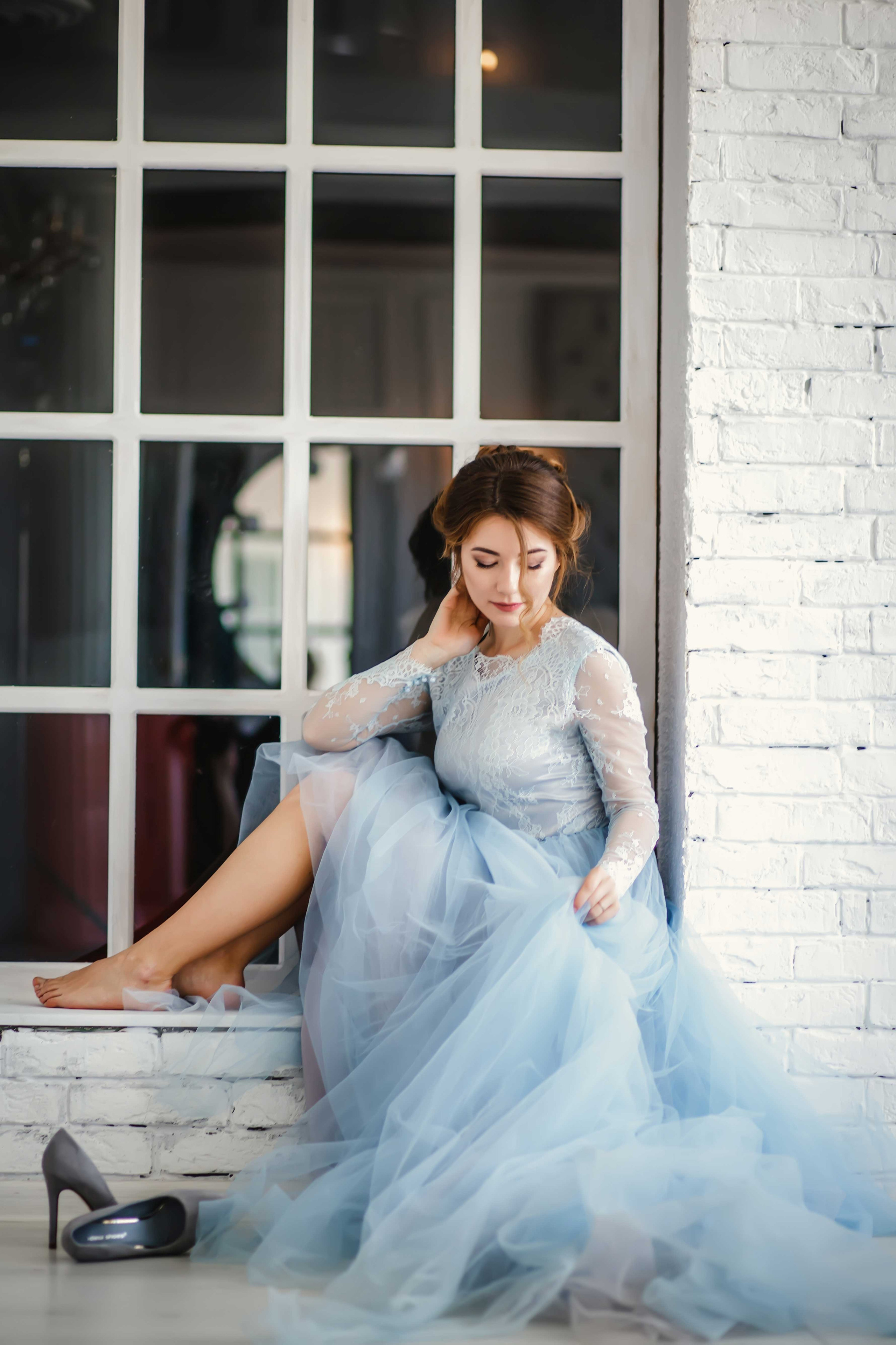 Нежная стильная невеста в голубом свадебном платье сидит подоконнике