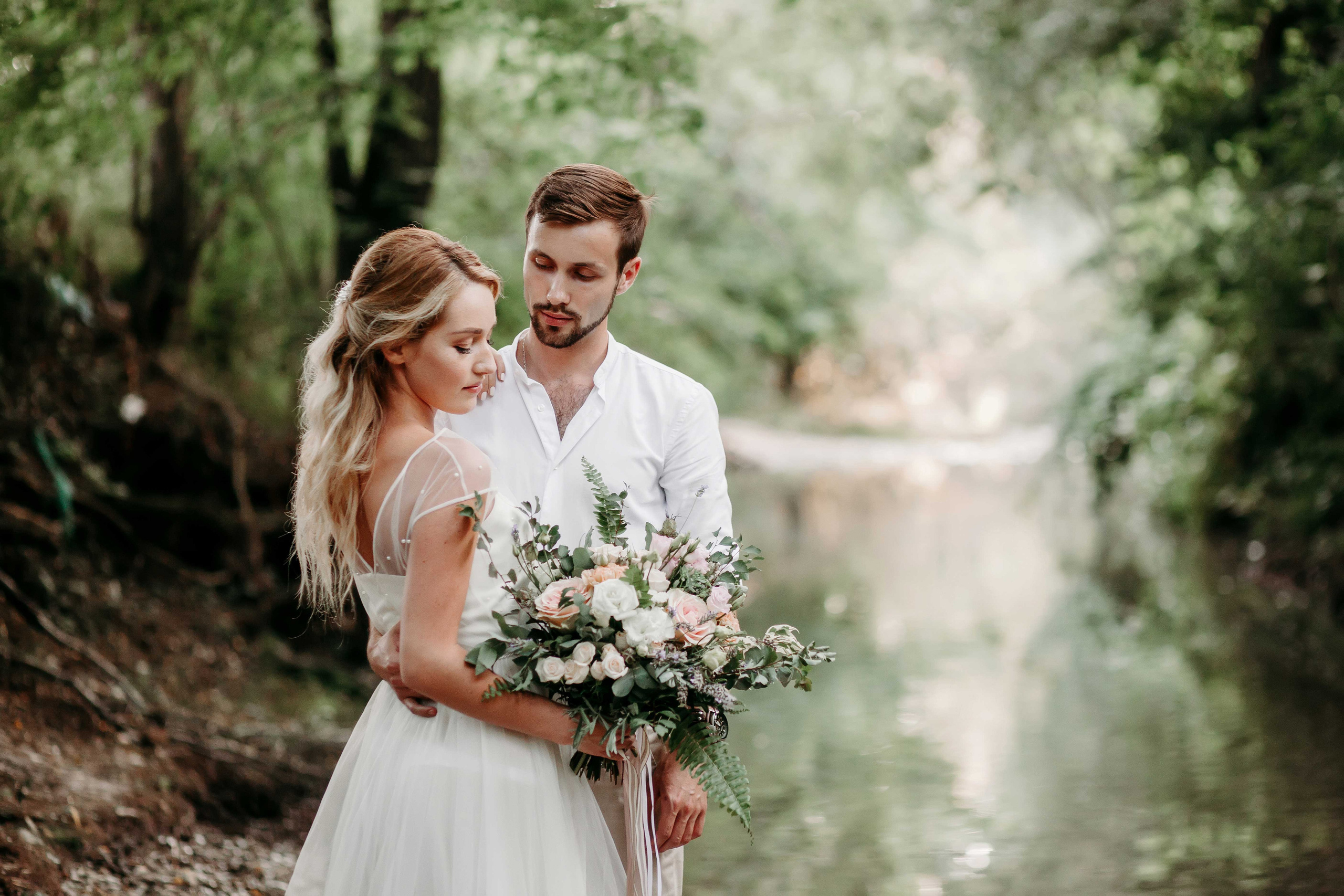 Невеста с распущенными светлыми волосами и жених в белой рубашке стоят на берегу реки взявшись за руки