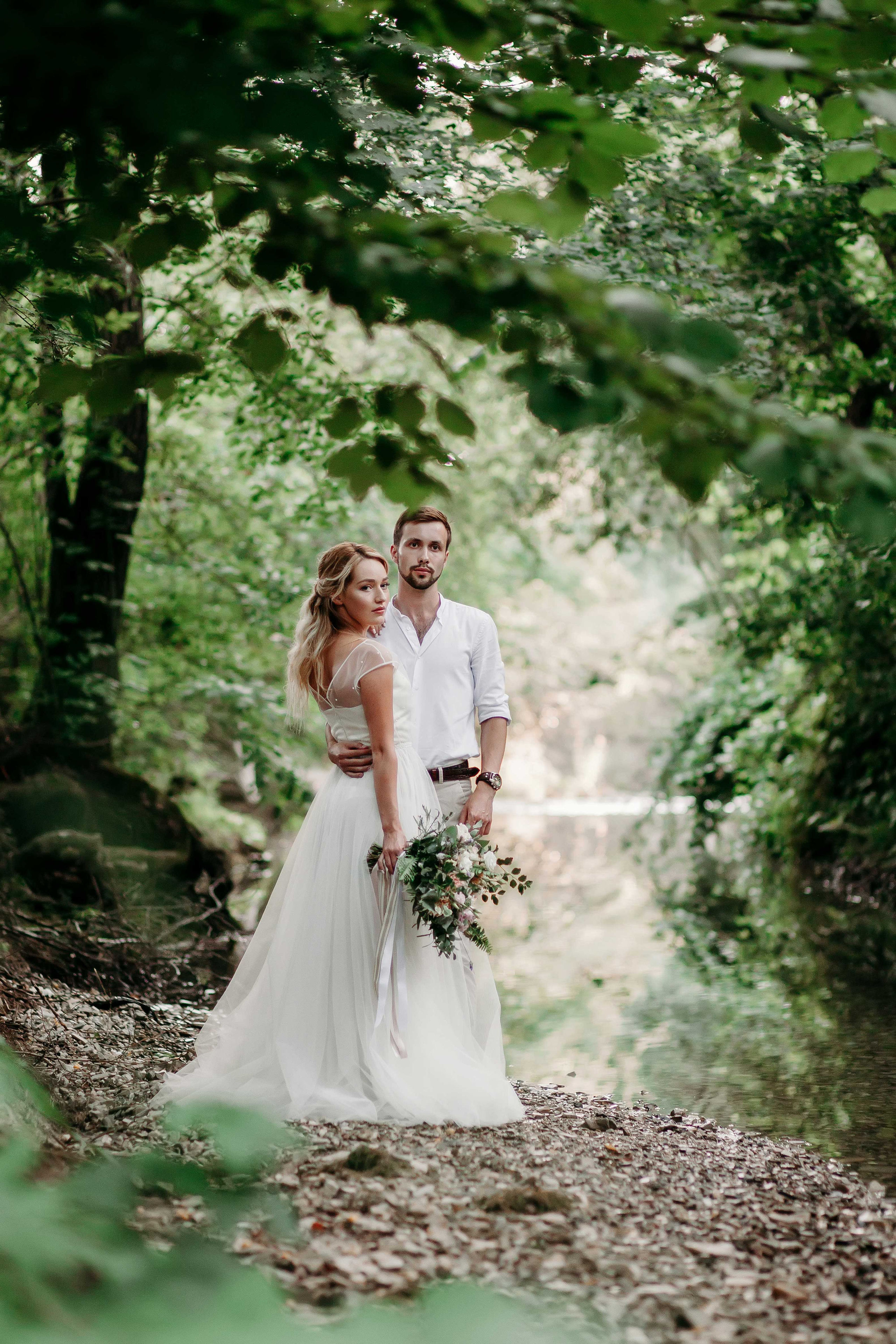 Невеста с распущенными светлыми волосами и жених в белой рубашке стоят на берегу реки обнявшись