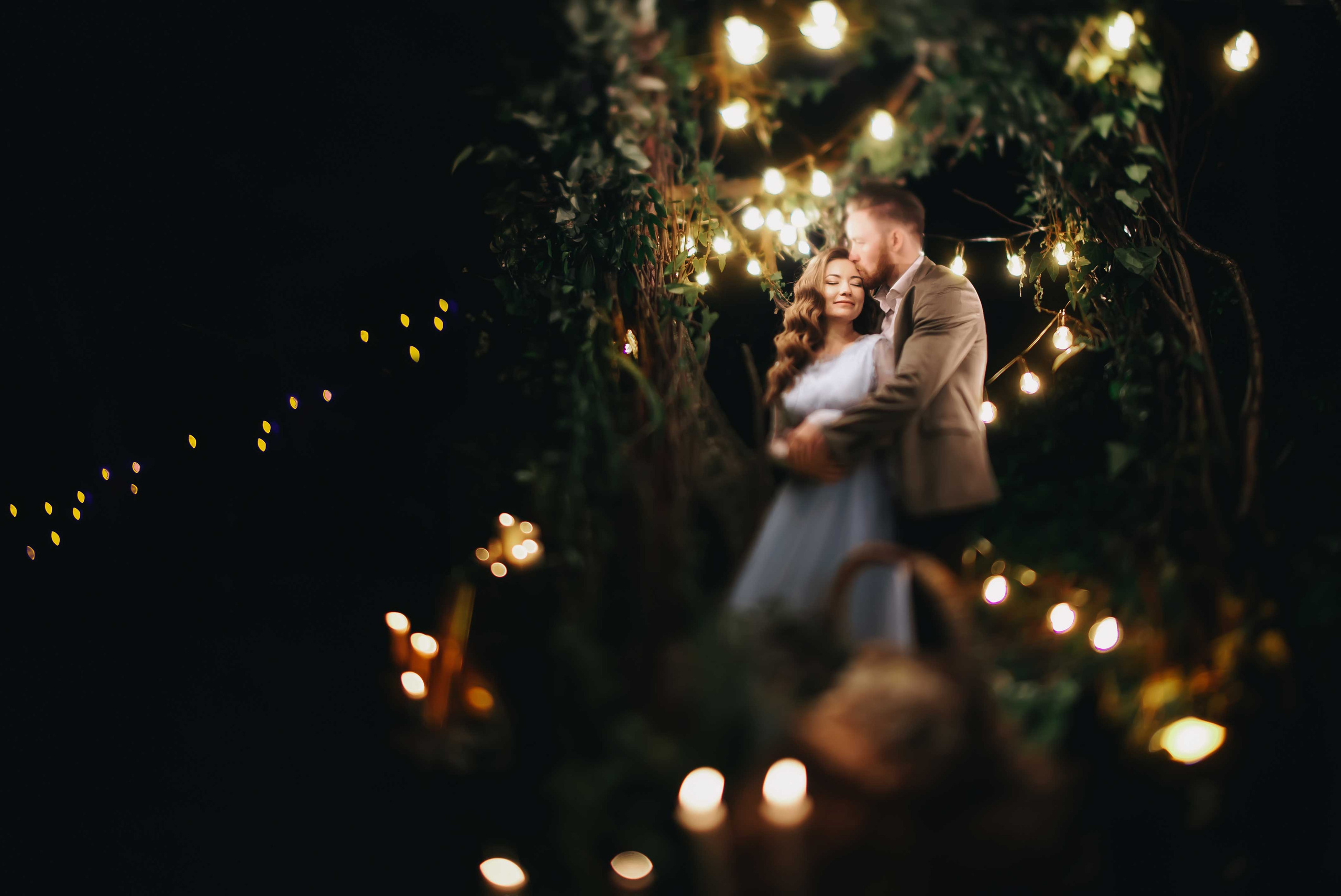 Жених обнимает невест в голубом свадебном платье с каштановыми распущенными волосами ночью в лесу украшенном гирляндами