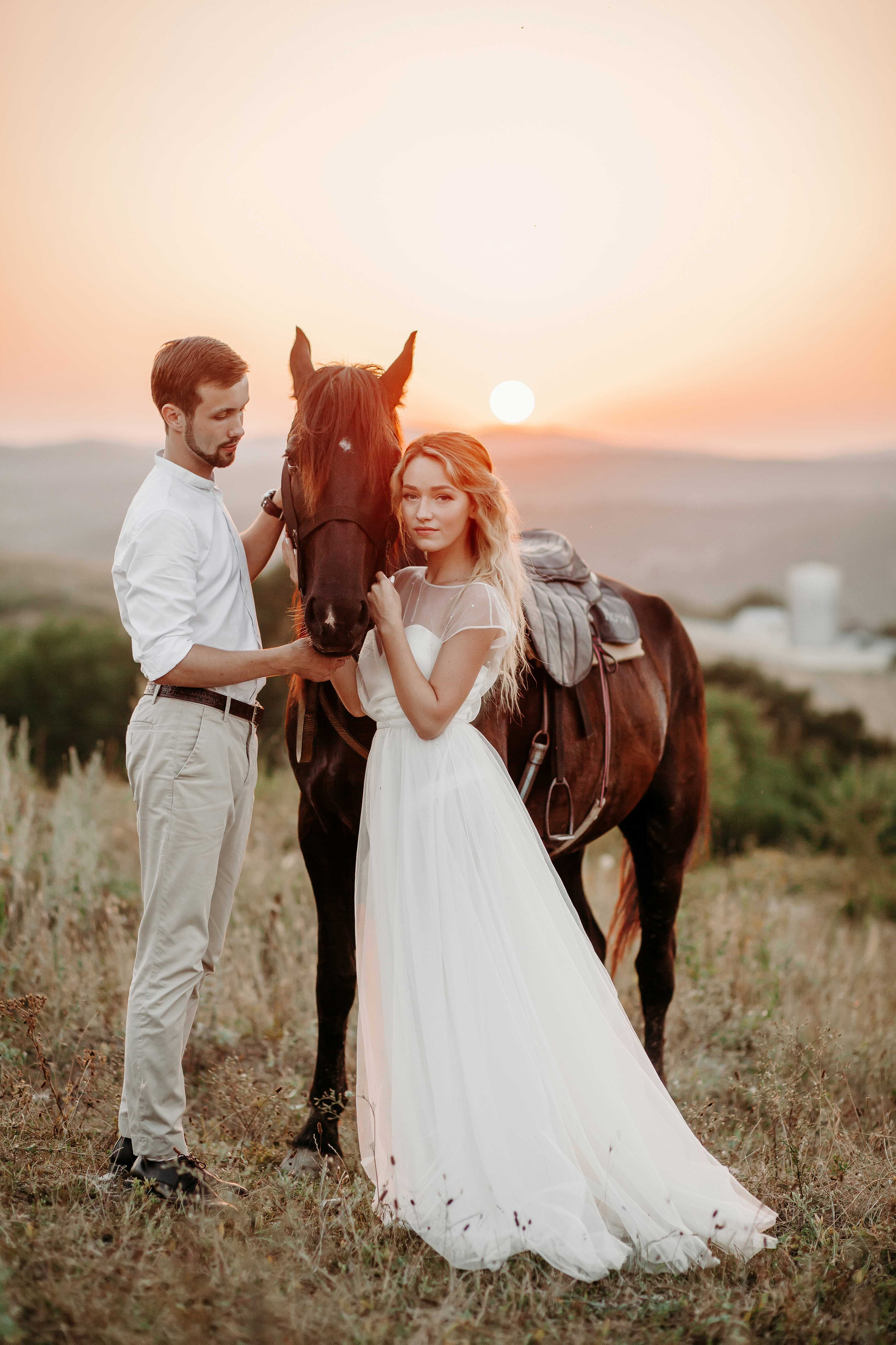 Блондинка невеста в белом платье и жених в бежевом стоят рядом с лошадью