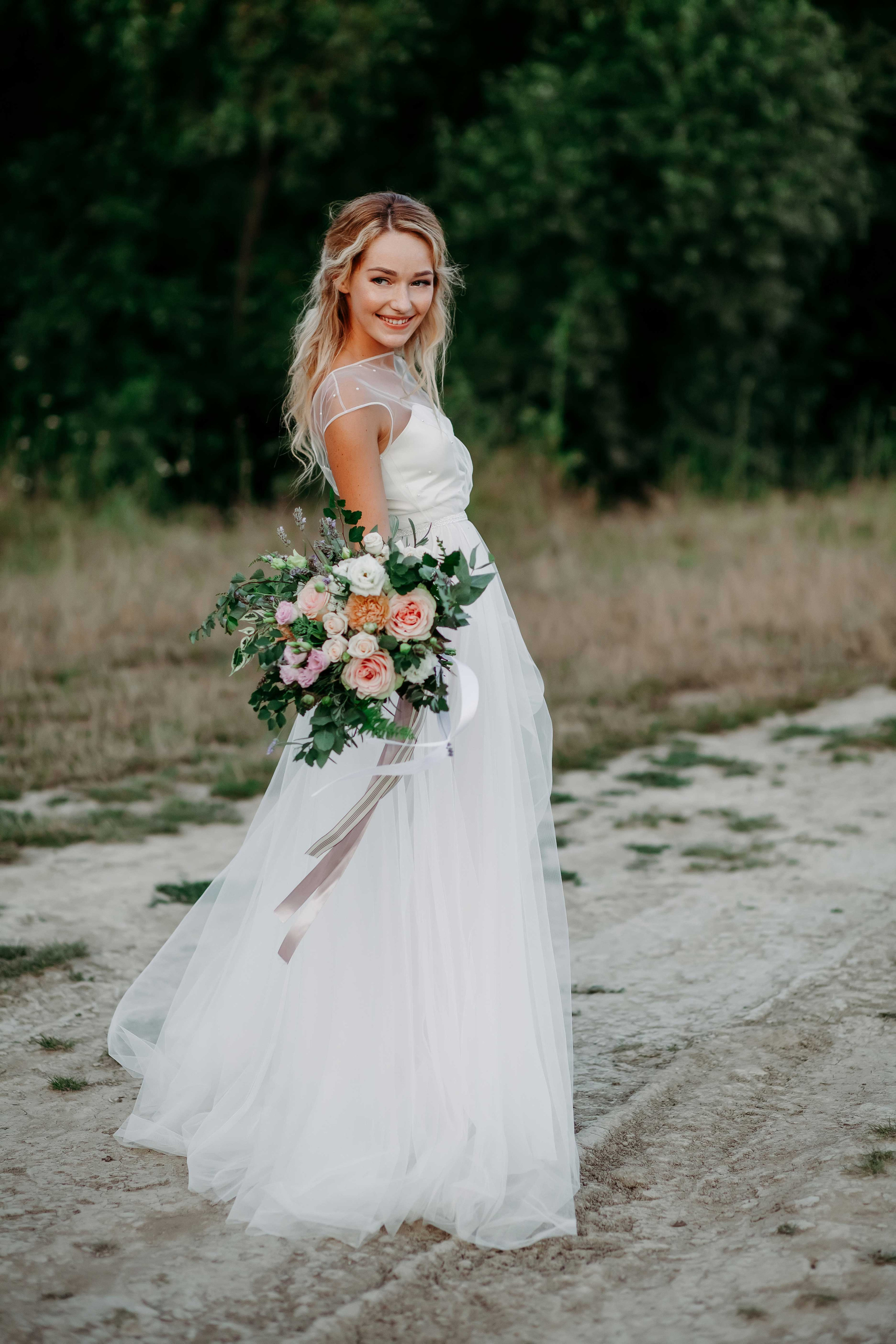 Невеста с распущенными светлыми волосами идет по тропинке со свадебным букетом в руках