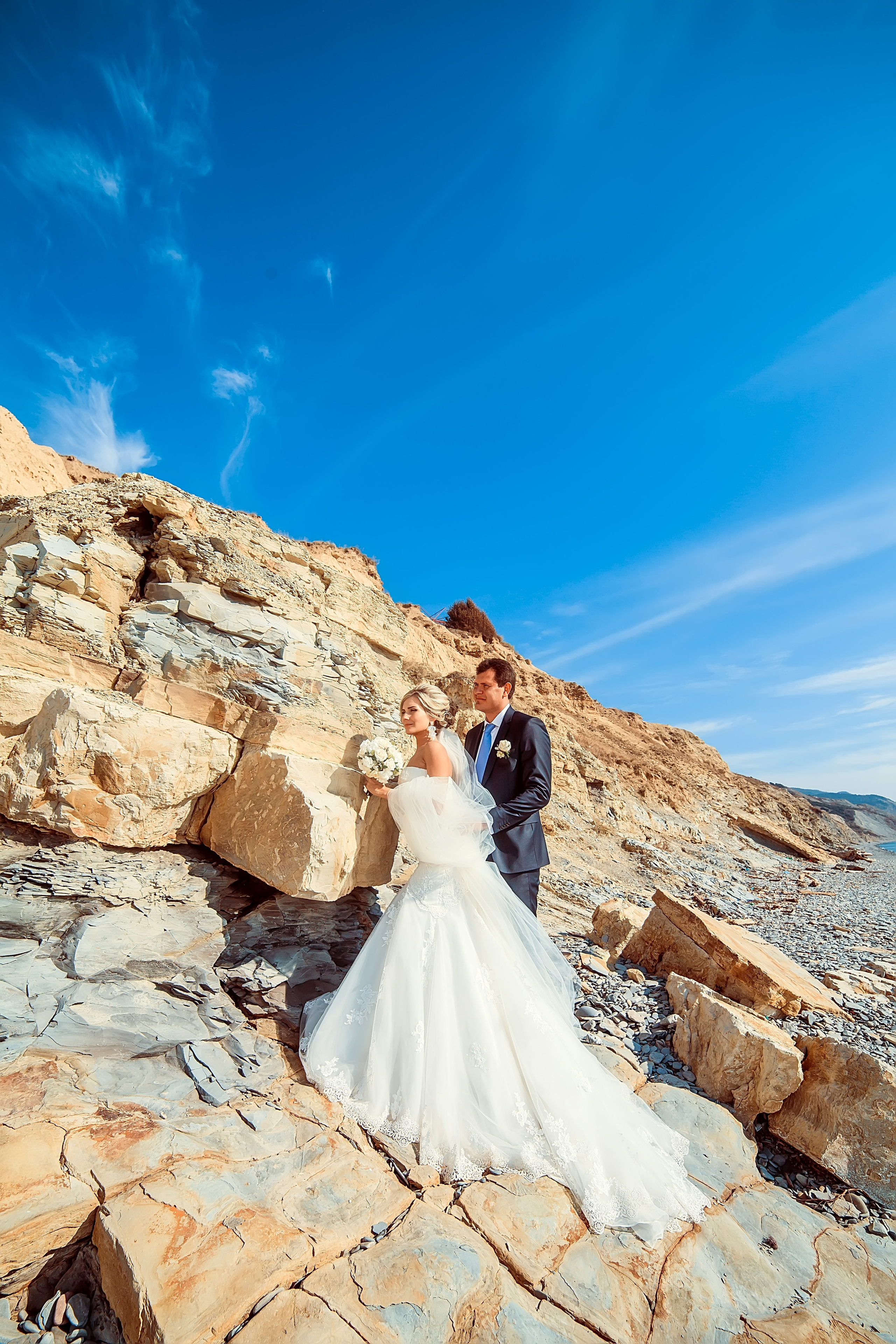 Жених и невеста стоят на фоне моря и скал обнявшись