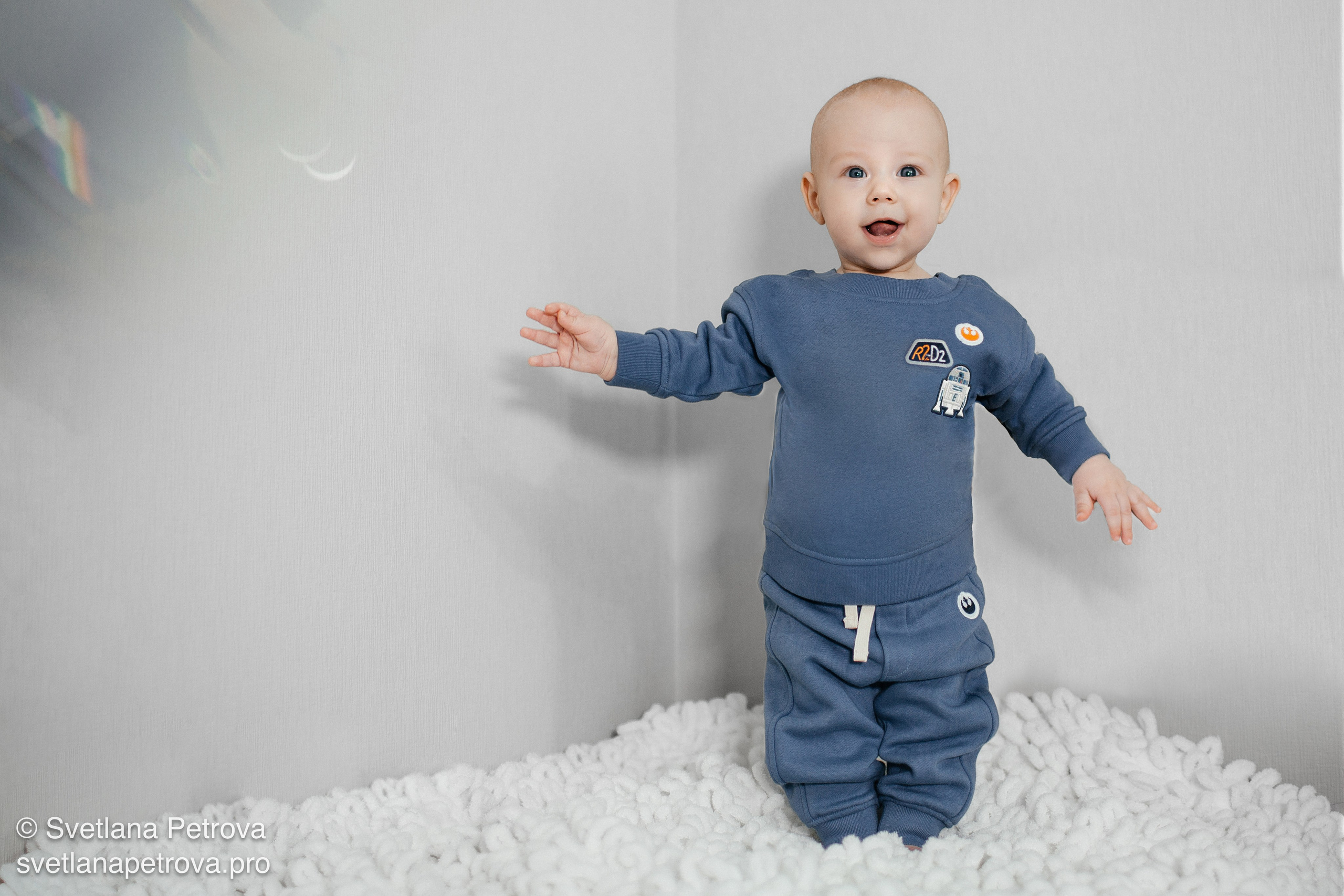 Малыш в голубом костюме на пушистом ковре — студийный кадр. Радостный младенец тянется к свету в студии. Студия, улыбающийся малыш, мягкий белый ковёр. В уютном костюме изучает мир вокруг. Пушистый ковёр, любопытный малыш, светлый фон. Счастливый малыш на ковре — семейная фотосессия. Студия, младенец в голубом, атмосфера уюта.