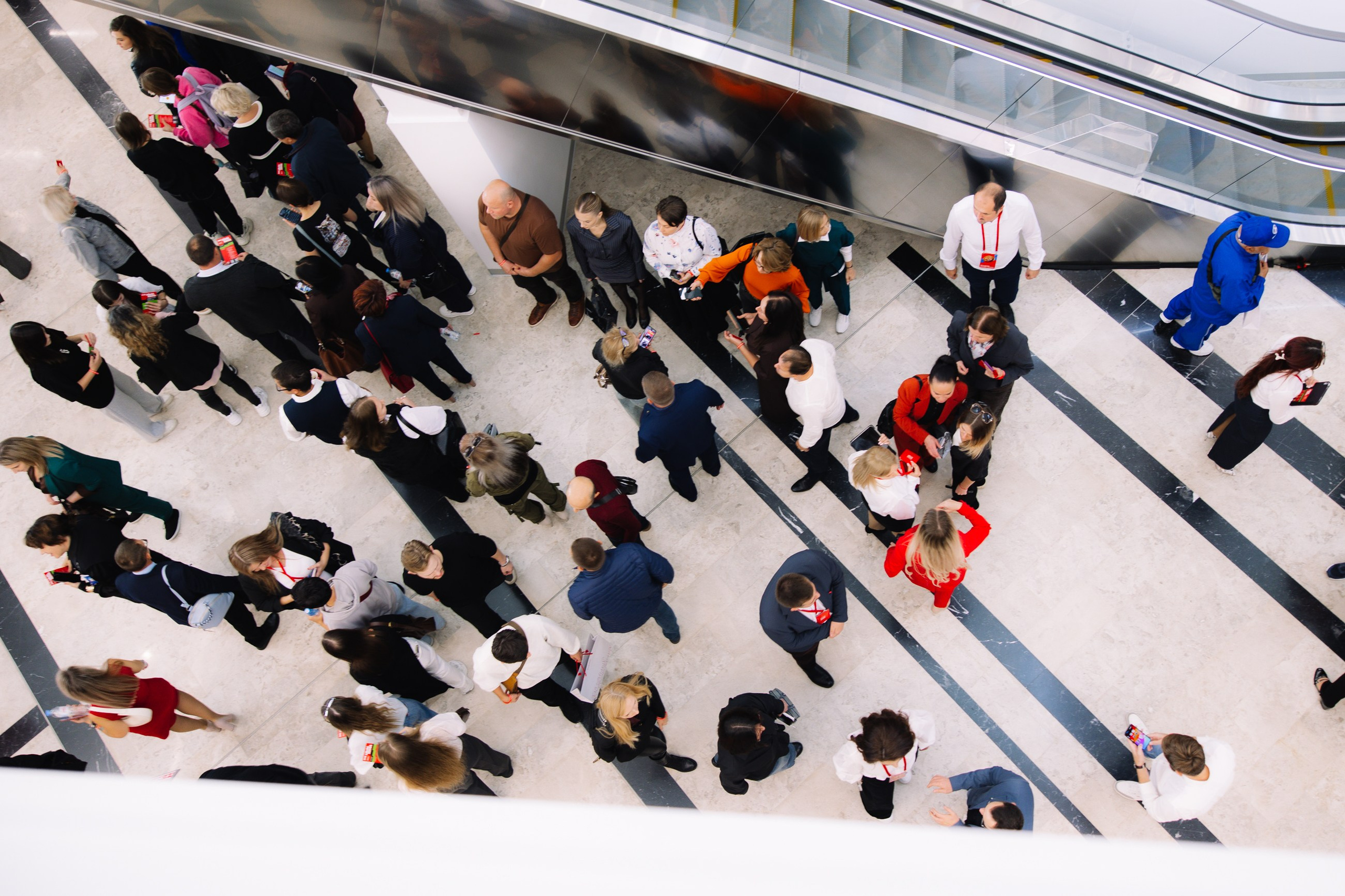 Вид сверху (ракурс): Оригинальный кадр участников в фойе, снятый вертикально вниз, ритм и движение толпы.