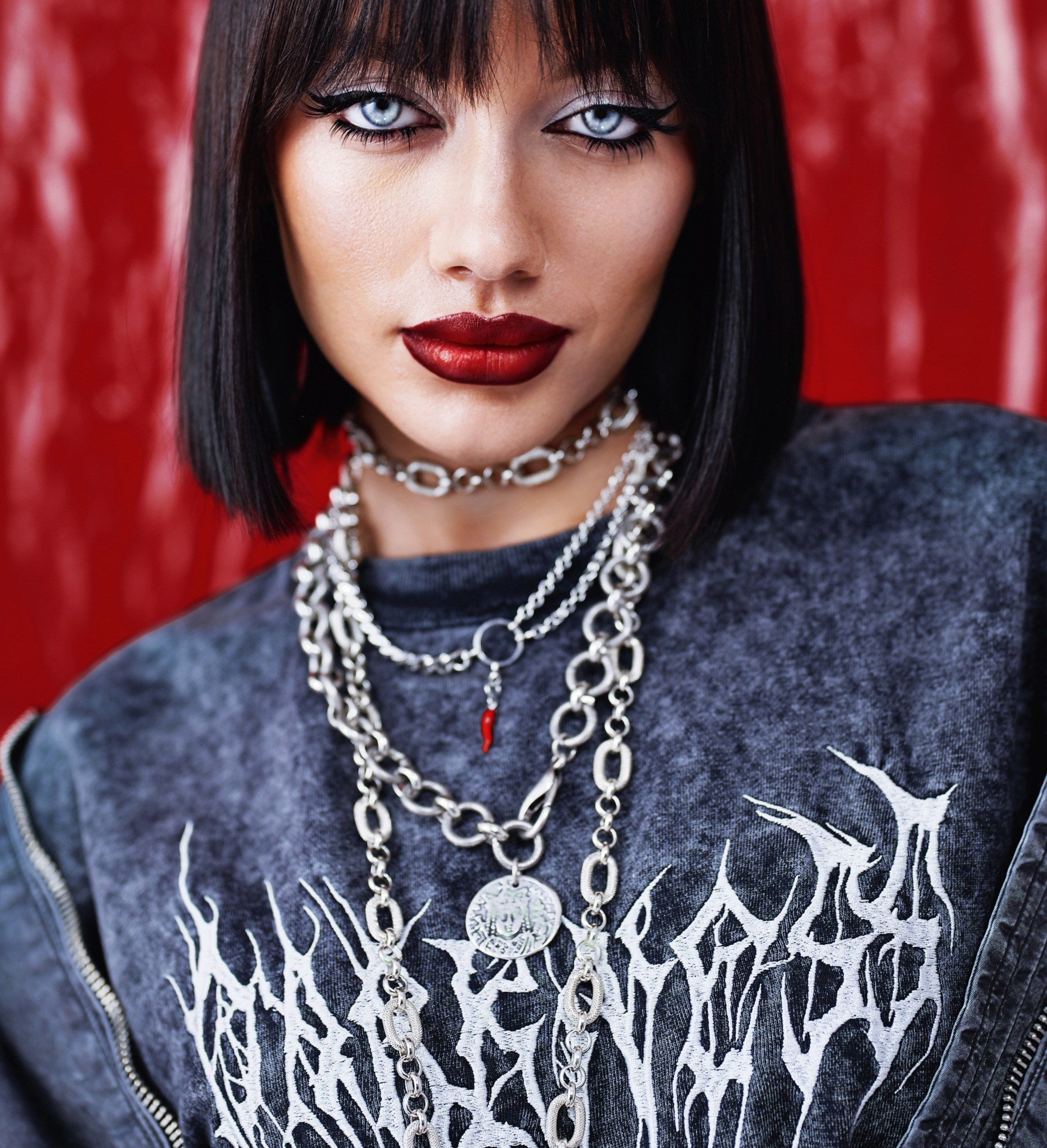 Портрет девушки с ярким макияжем, темными волосами и металлическими украшениями на шее, крупным планом на фоне красной глянцевой поверхности