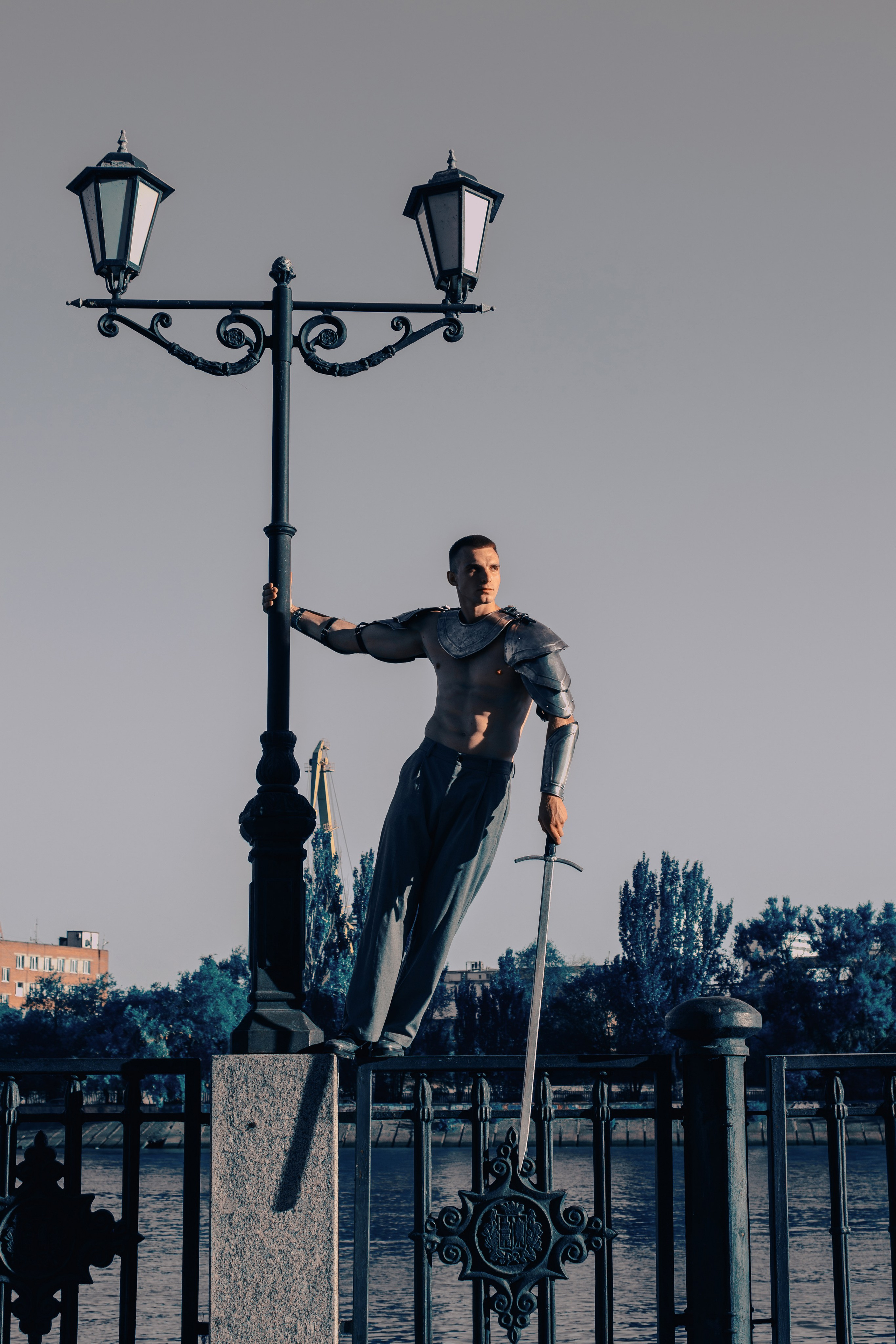 В полный рост в доспехах, брюках и туфлях стоит на заборе мужчина с мечом в руках