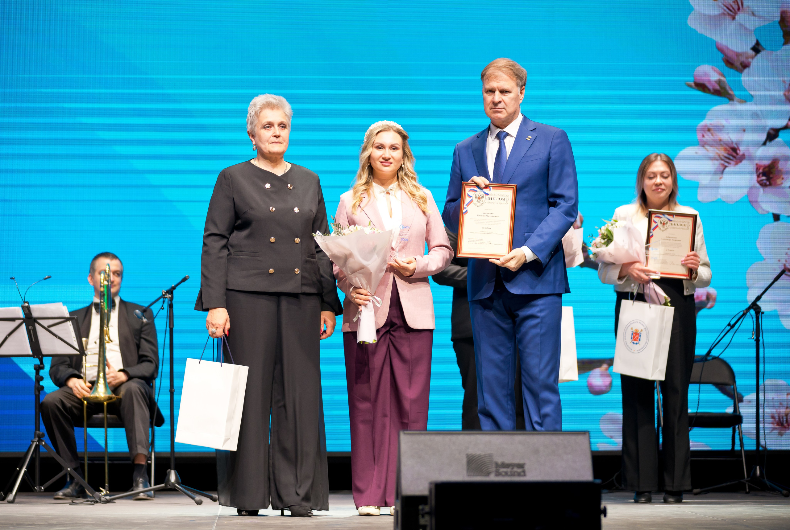 Церемония награждения Дня местного самоуправления. Женщина в чёрном костюме, лауреат в розовом пиджаке и глава администрации с грамотой на сцене | Фото: Евгений Мальцев.