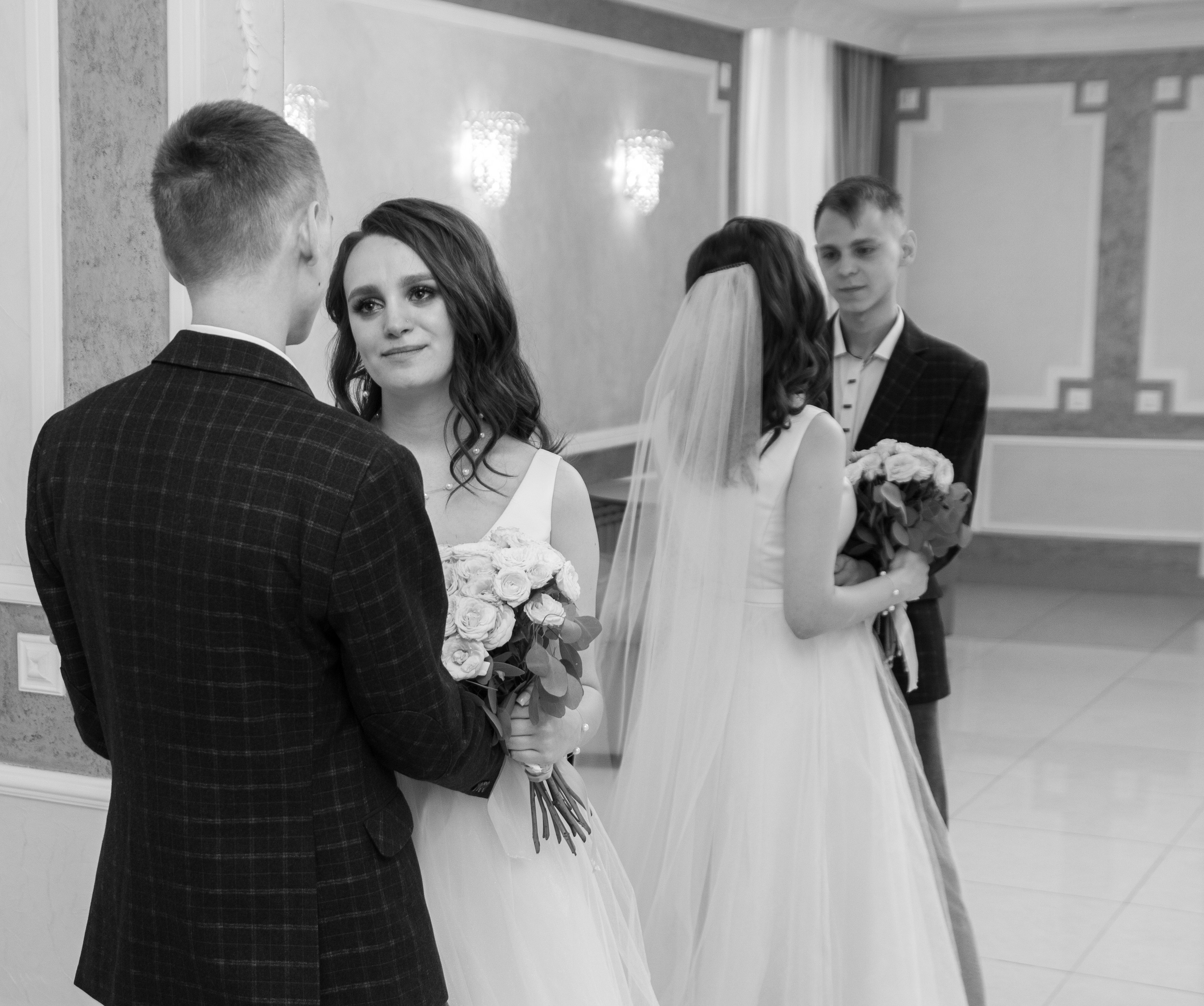 Жених и невеста стоят лицом друг к другу в зале, невеста держит букет.