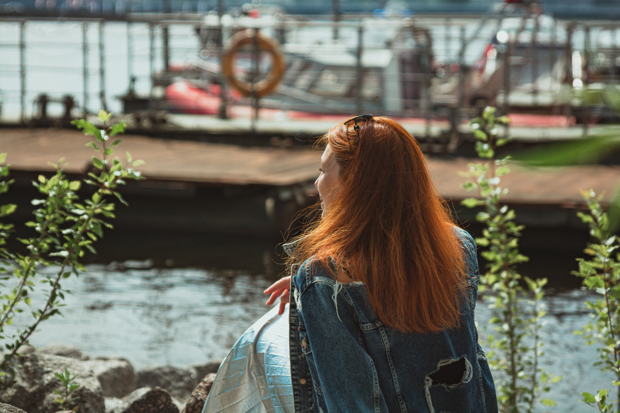Девушка сидит на берегу у причала в джинсовой куртке