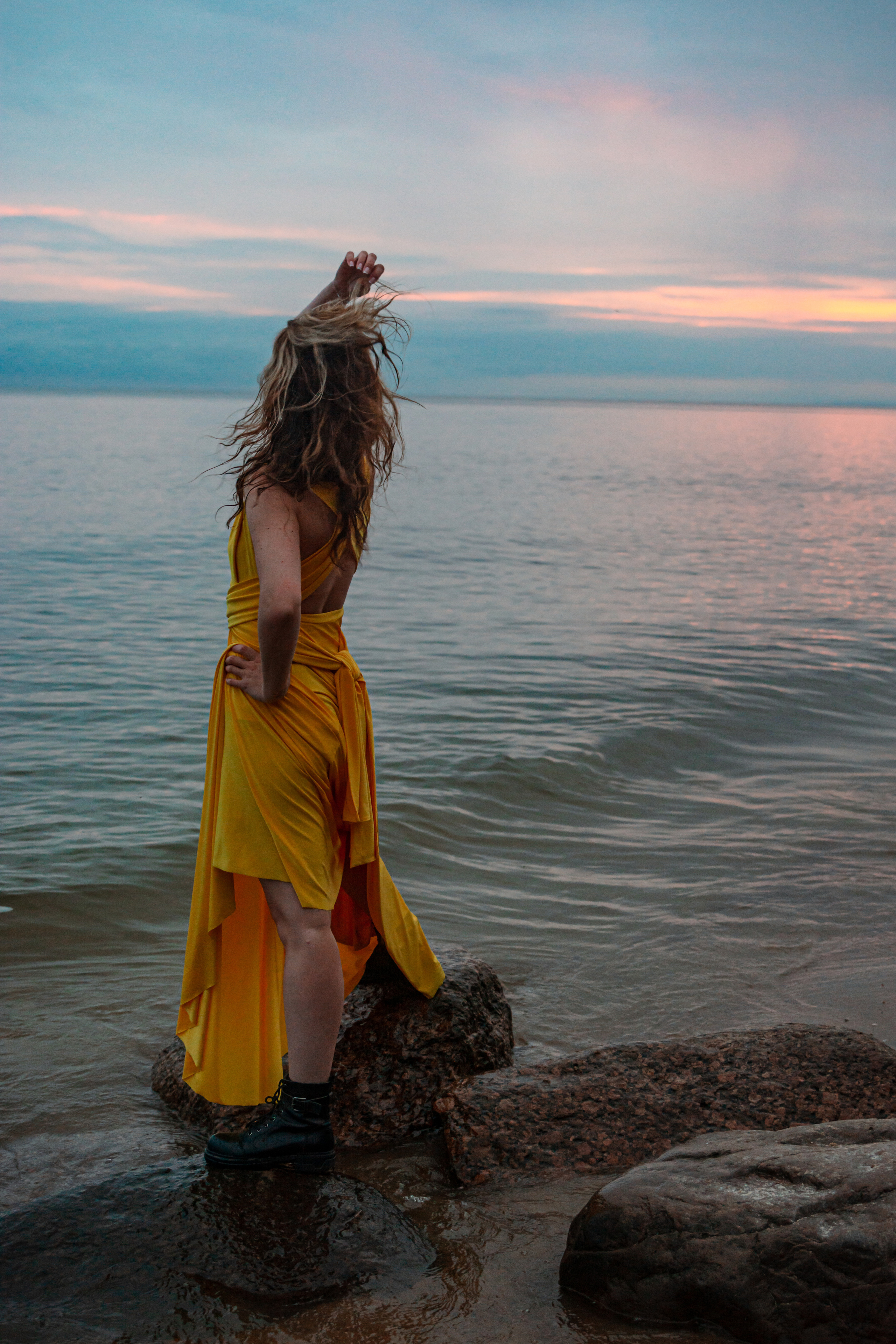 Девушка в желтом платье с улыбкой на лице позирует во время фотосессии на берегу Финского залива, где закат создает прекрасное освещение.